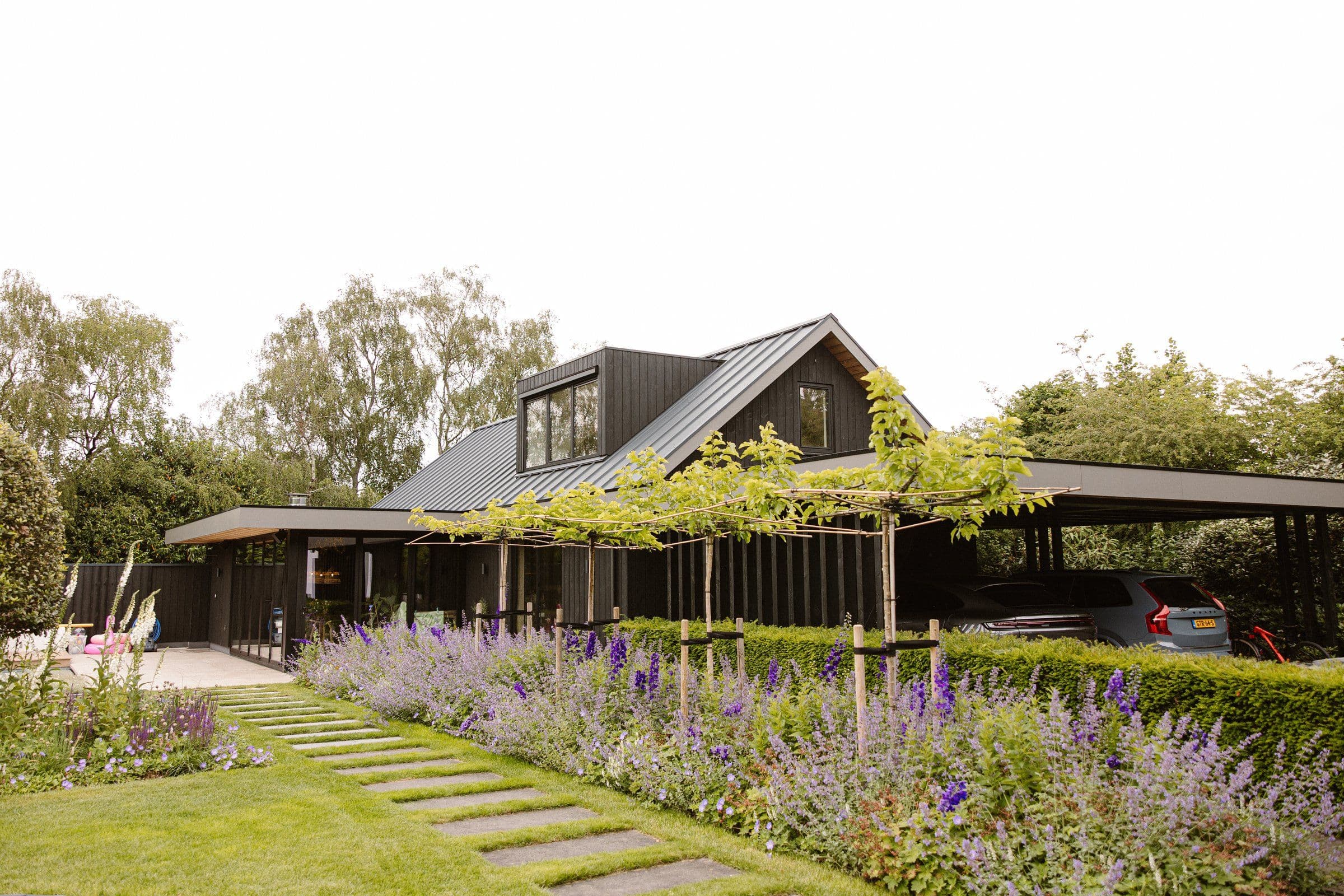 Moderne woning met zwarte houten gevel en carport, omgeven door een groene tuin met paadje en bloemen.