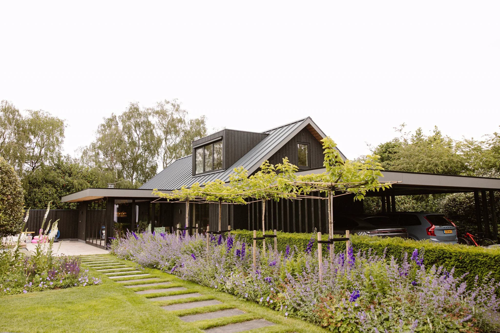 Moderne woning met zwarte houten gevel en carport, omgeven door een groene tuin met paadje en bloemen.