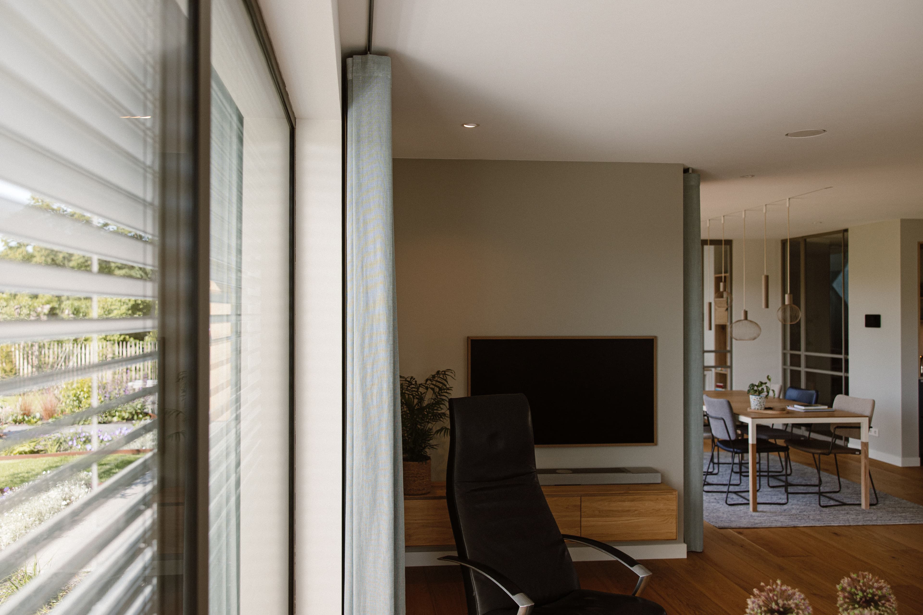 Moderne woonkamer met een zwarte stoel voor een houten meubel met televisie, en in de achtergrond een eettafel met stoelen en hanglampen