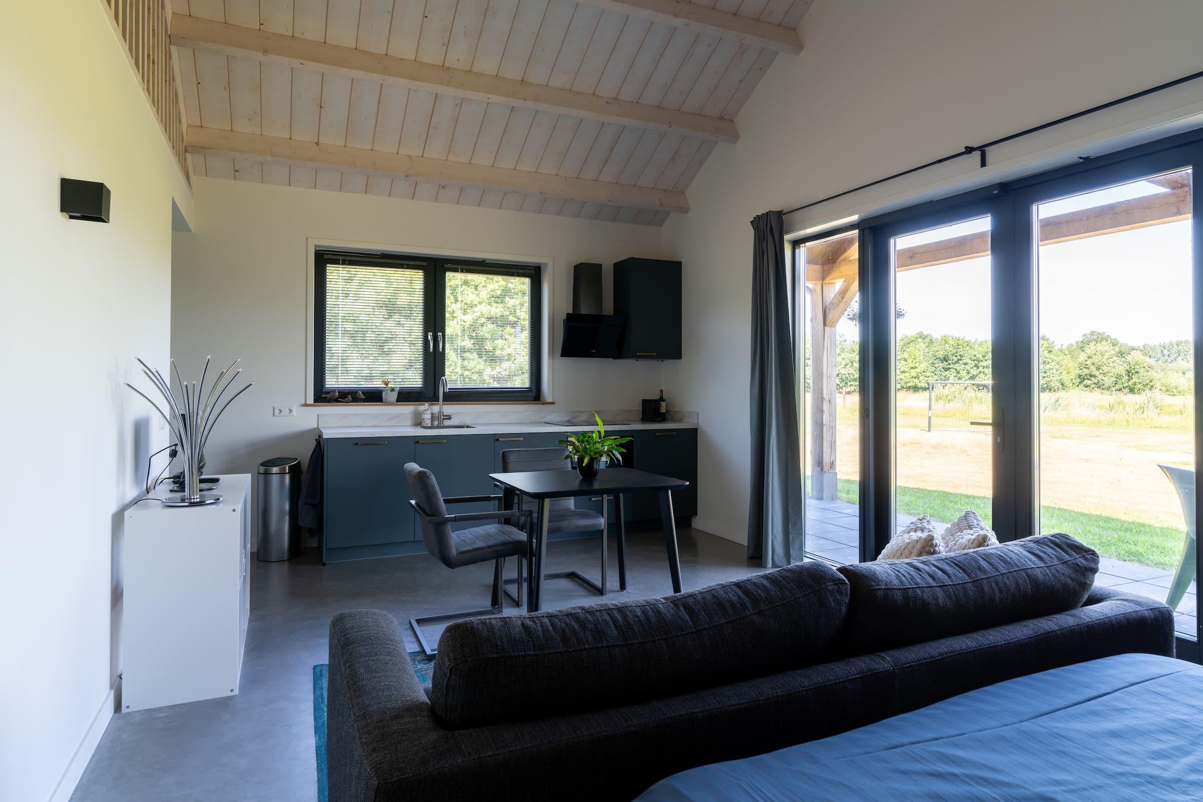 Woonkamer met zwarte bank, zwarte eettafel en stoelen, modern keukenblok, grote ramen en uitzicht op tuin