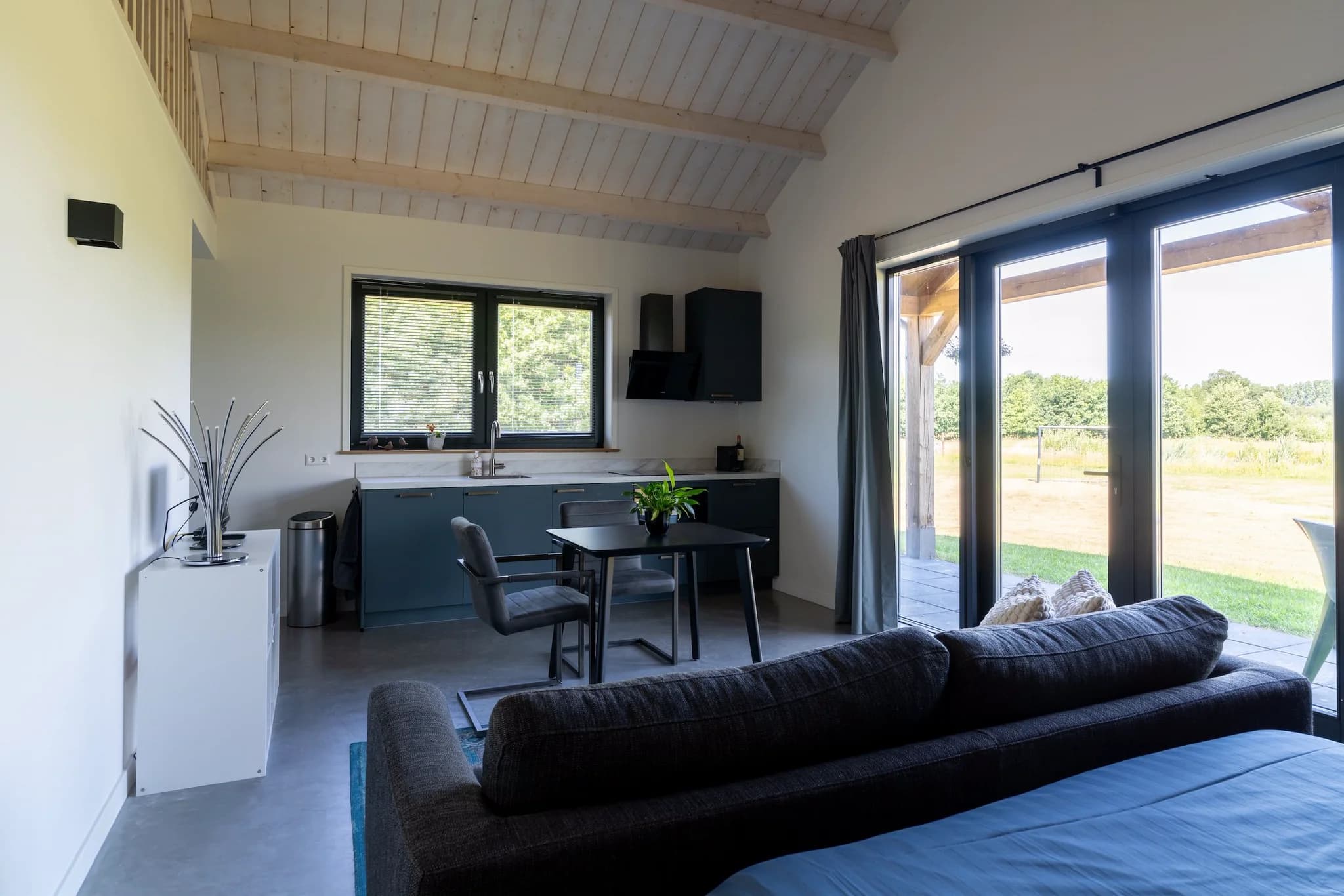 Woonkamer met zwarte bank, zwarte eettafel en stoelen, modern keukenblok, grote ramen en uitzicht op tuin