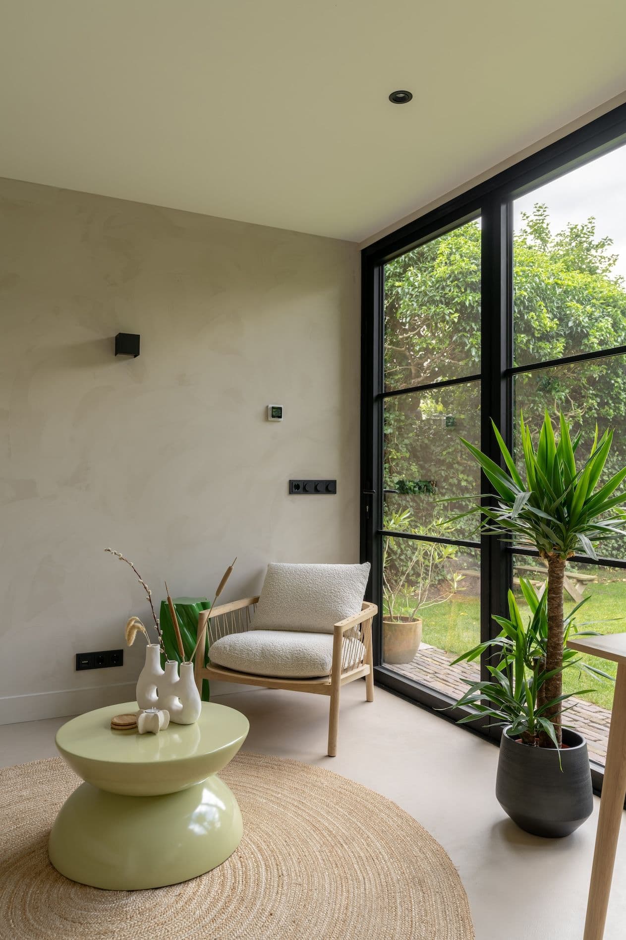 Zithoek met een houten stoel met witte bekleding, groene salontafel, planten en grote ramen met uitzicht op tuin.