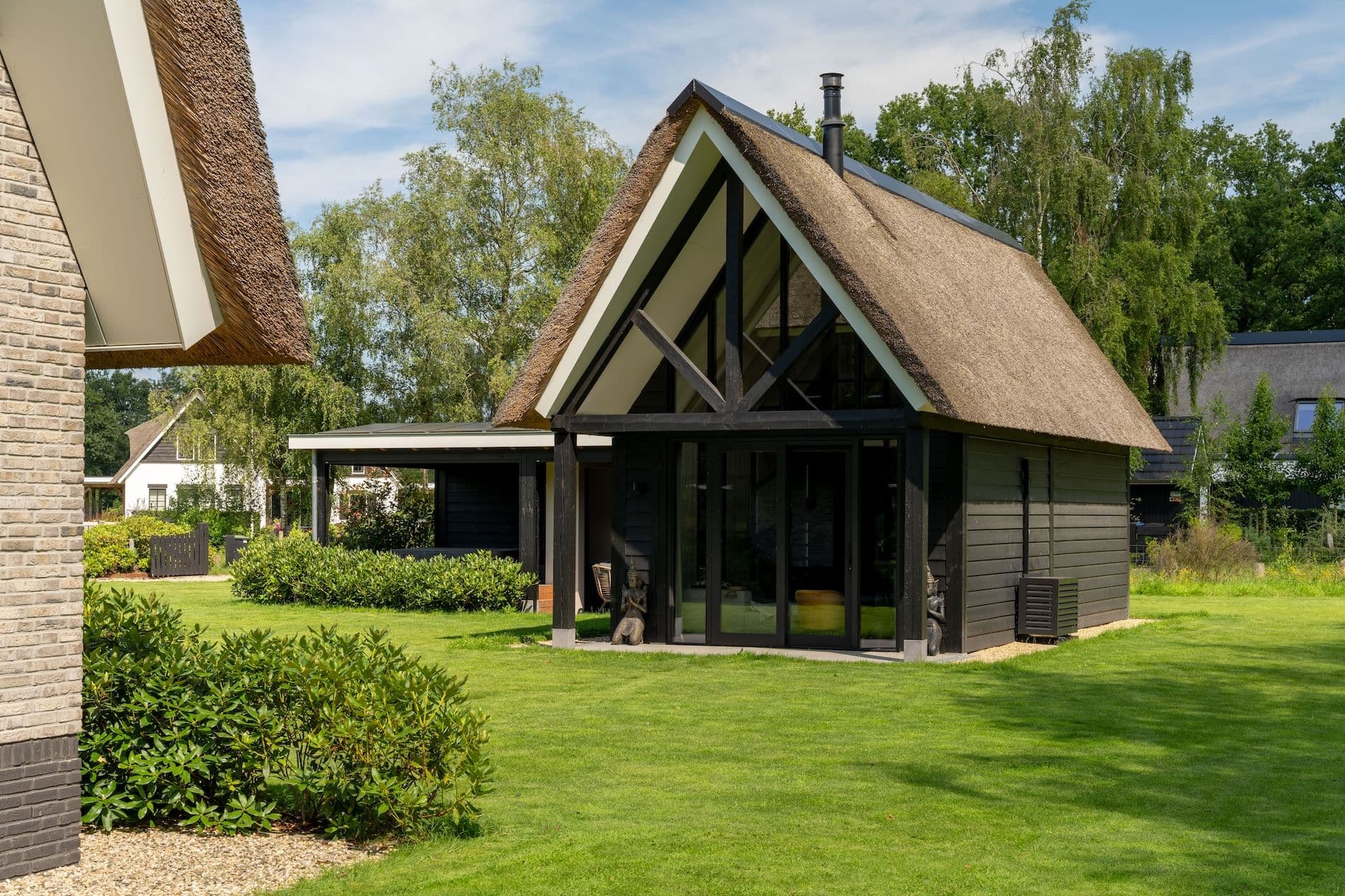Zwarte houten vakantiewoning met rieten dak en grote glazen schuifdeuren, omgeven door groene tuin met gras en struiken