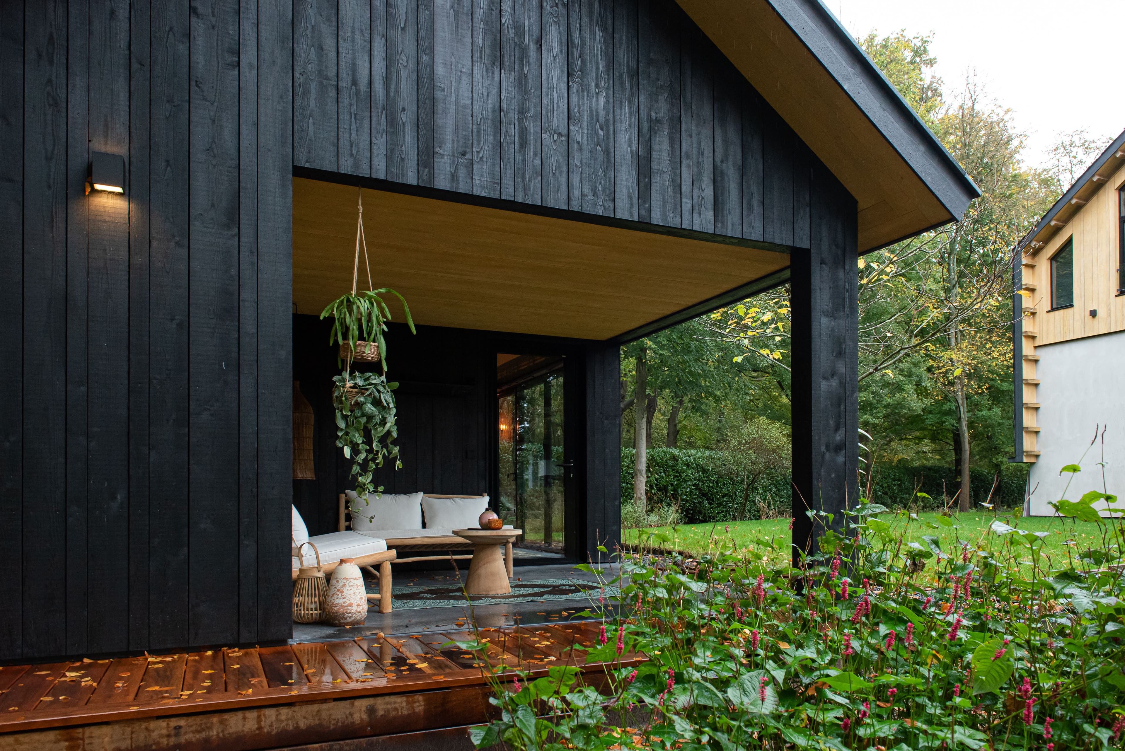 Zwarte houten veranda met bankje, tafel en hangplanten naast een groene tuin met rode bloemen
