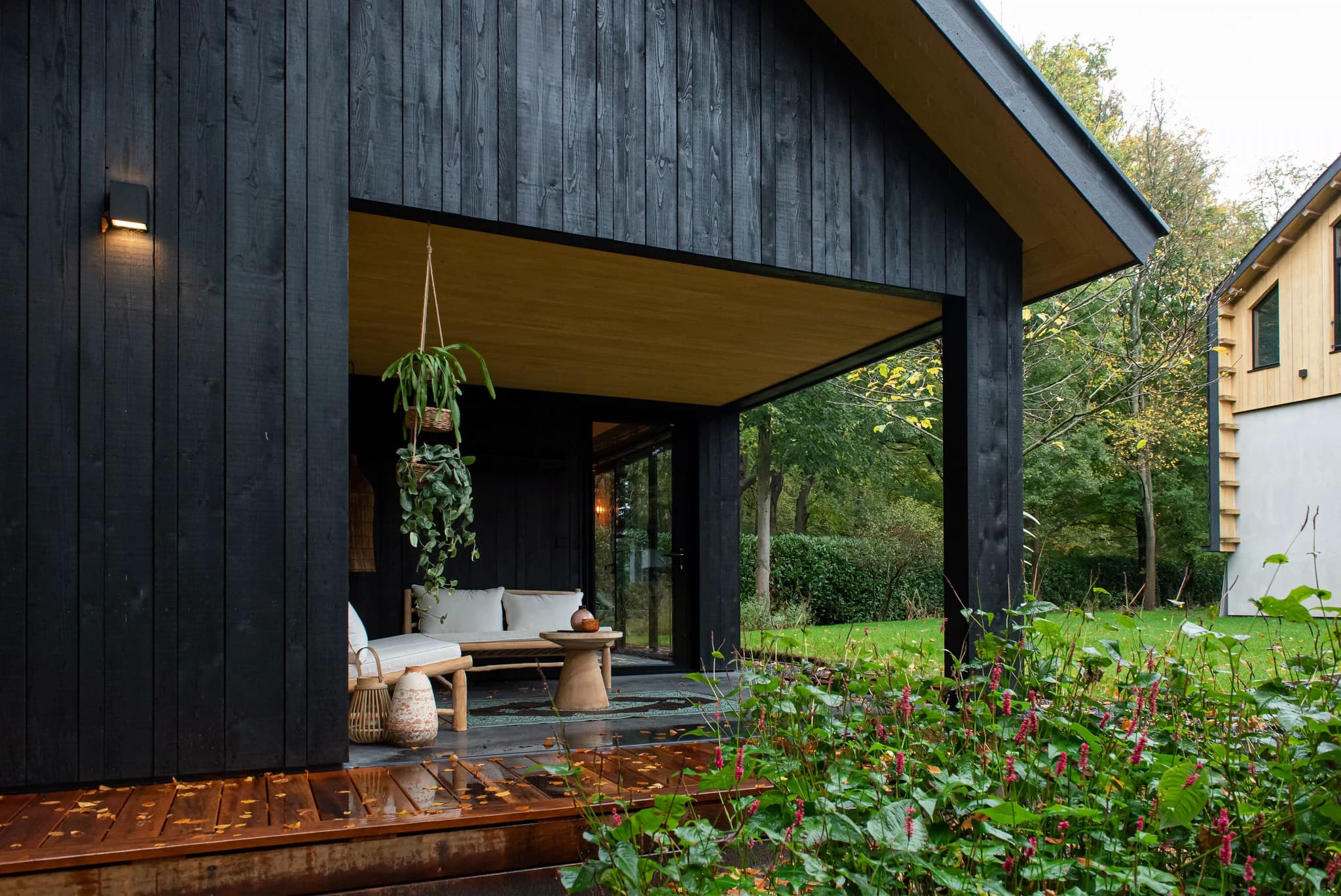 Zwarte houten veranda met bankje, tafel en hangplanten naast een groene tuin met rode bloemen