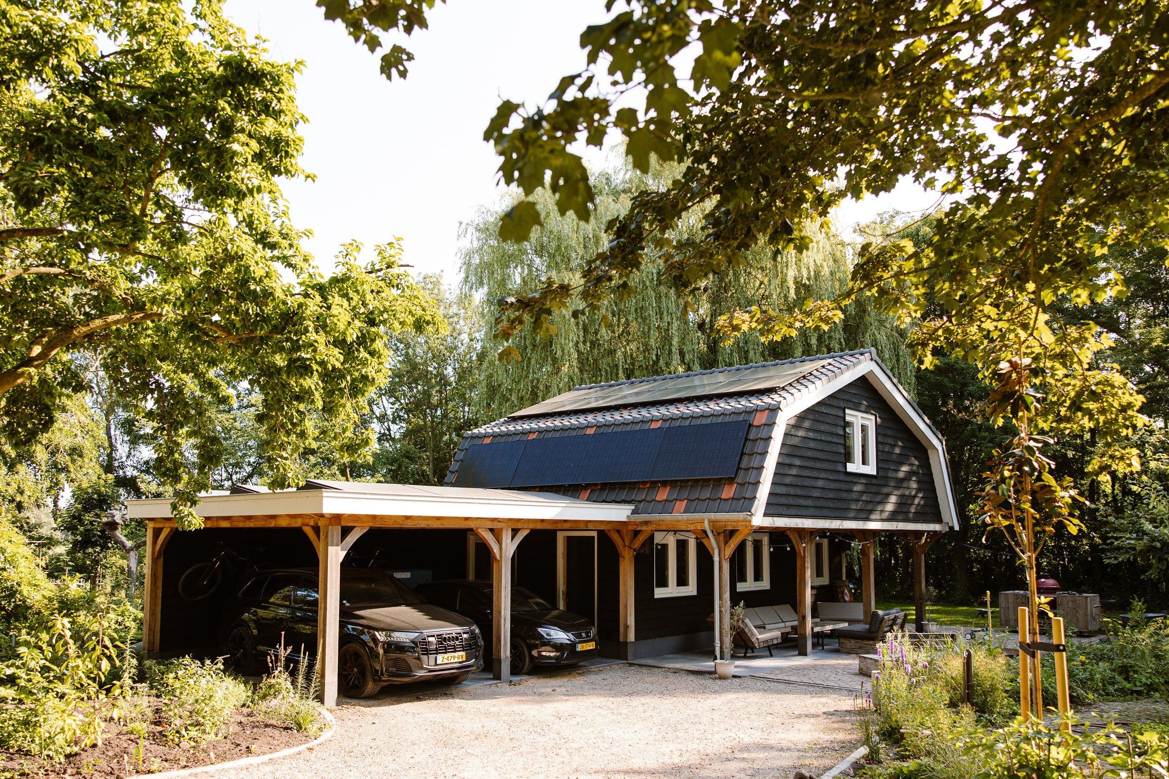Zwarte houten woning met zonnepanelen op het dak, twee auto's onder een houten carport en groene tuin met bomen.