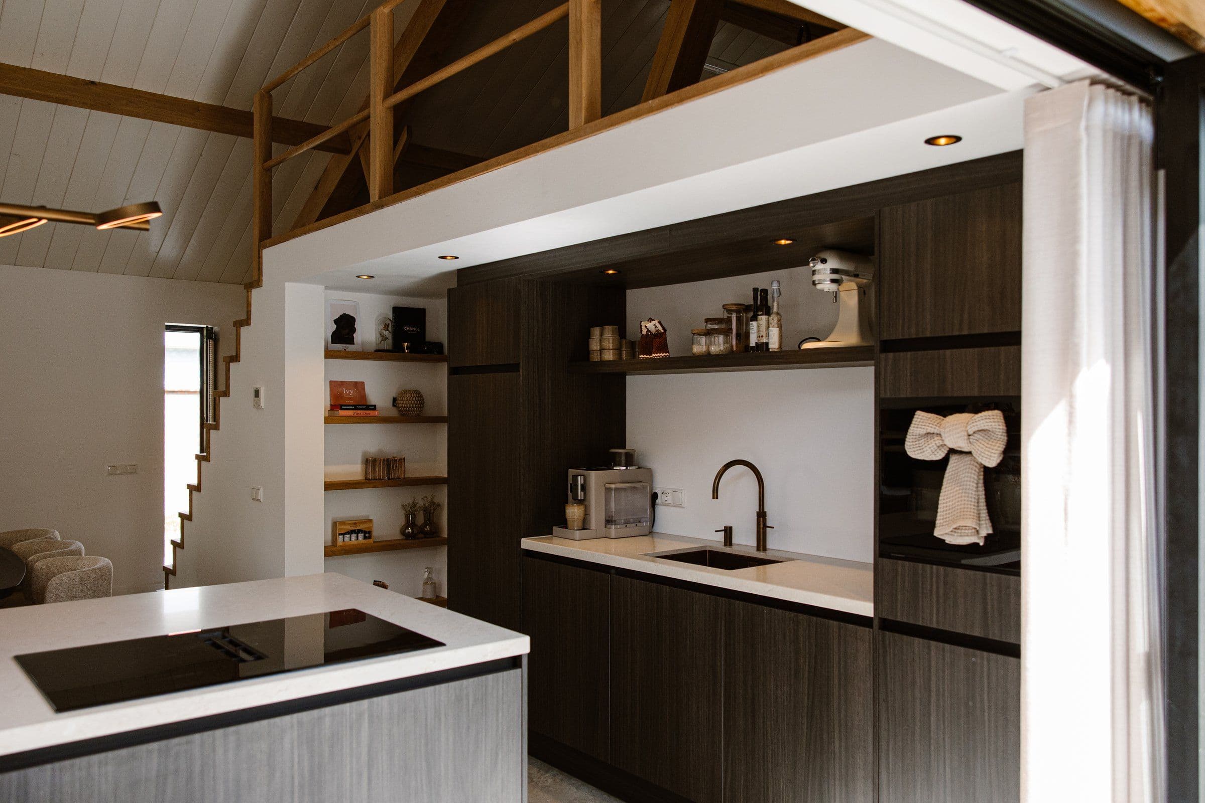 Binnenzicht van een moderne keuken met donkergrijze kasten, wit werkblad, ingebouwde spoelbak en open houten planken onder een houten mezzanine
