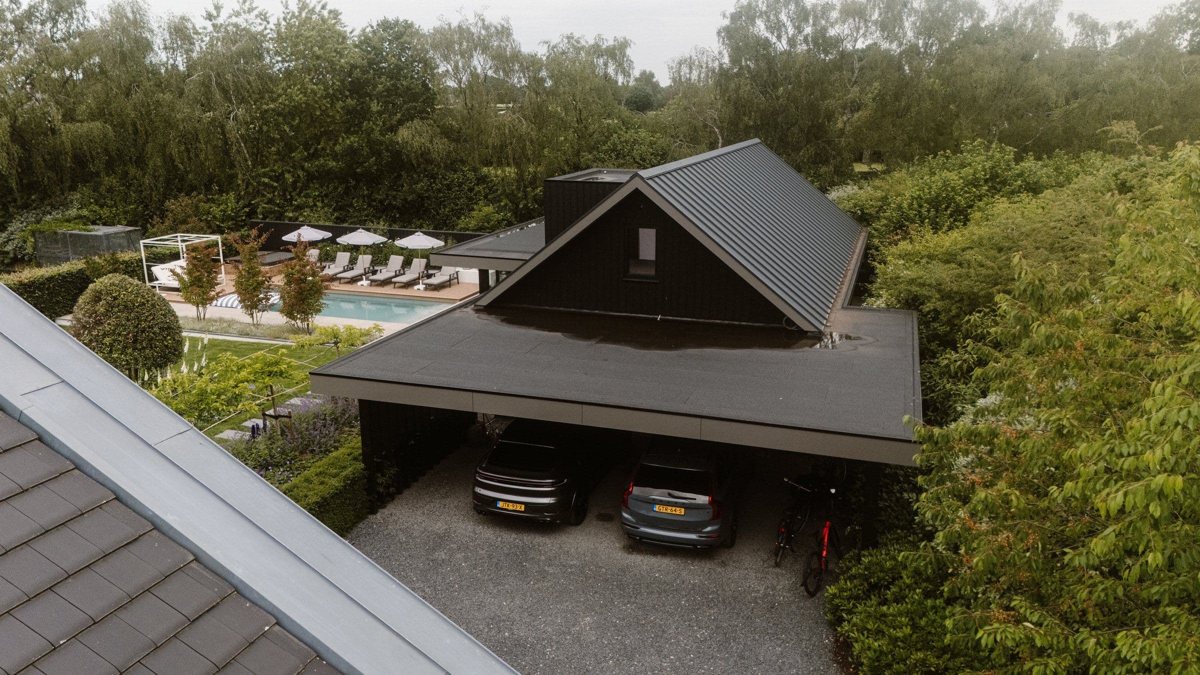 Luchtfoto van een moderne woning met donkere overkapping en twee auto's, zwembad met ligstoelen en parasols in de achtertuin, veel groen rondom
