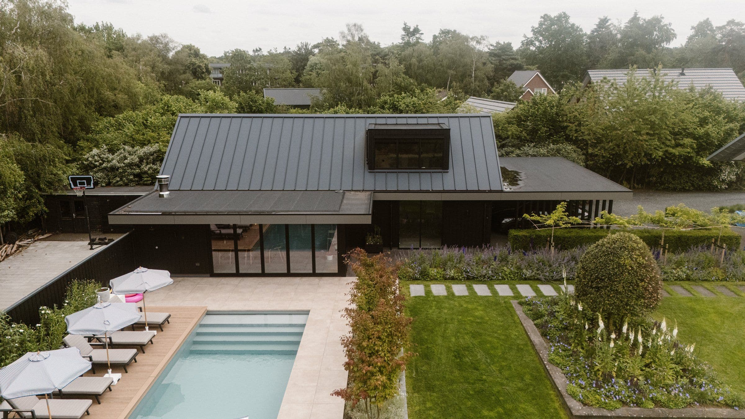 Luchtfoto van een moderne woning met donker schuin dak, terras met zwembad en meerdere ligbedden, omringende groene tuin en bomen