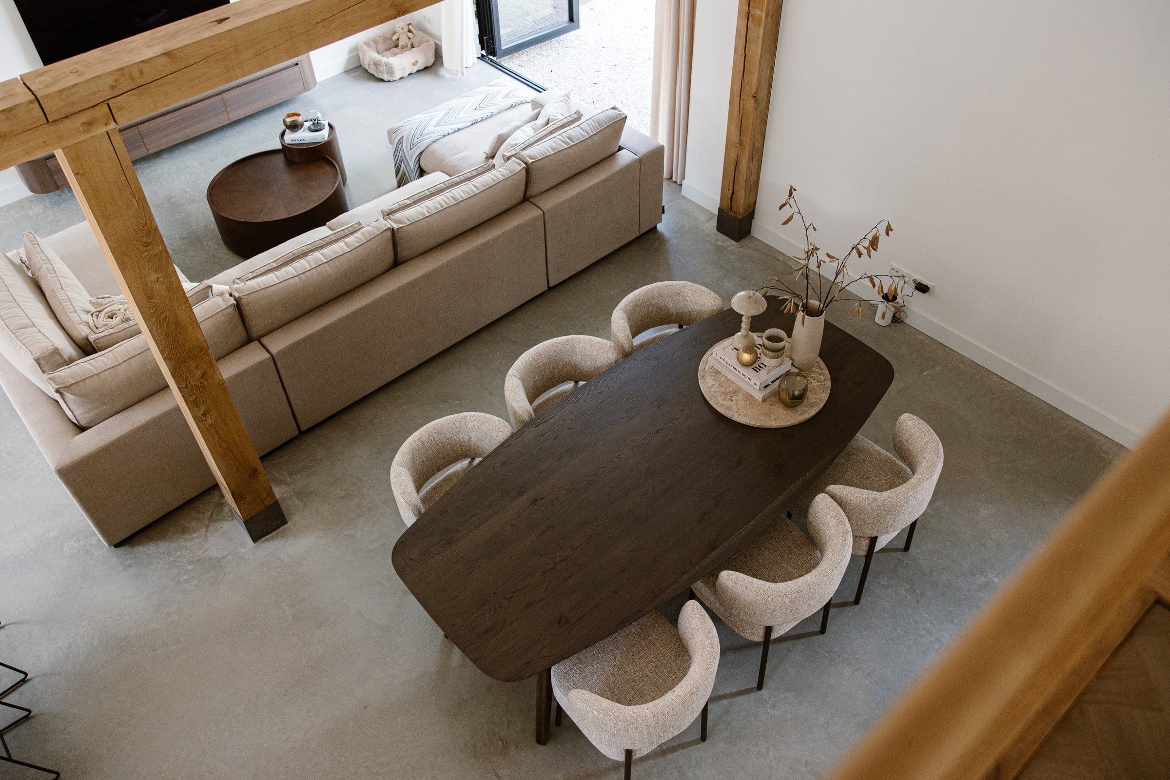 Woonkamer en eetkamer met een grote beige bank, een houten eettafel en zes crèmekleurige stoelen, decoratie op tafel en zicht op buiten.