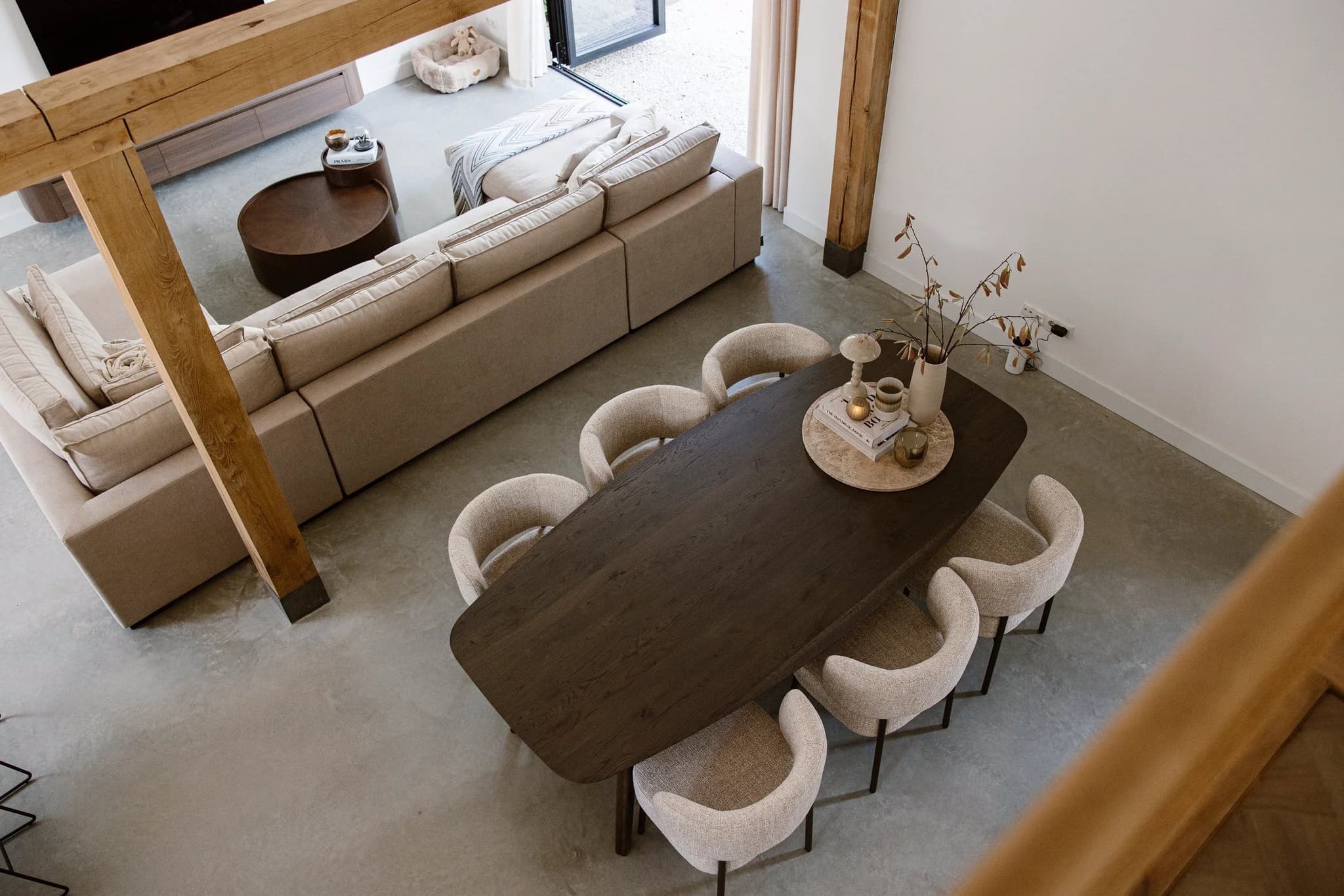 Woonkamer en eetkamer met een grote beige bank, een houten eettafel en zes crèmekleurige stoelen, decoratie op tafel en zicht op buiten.