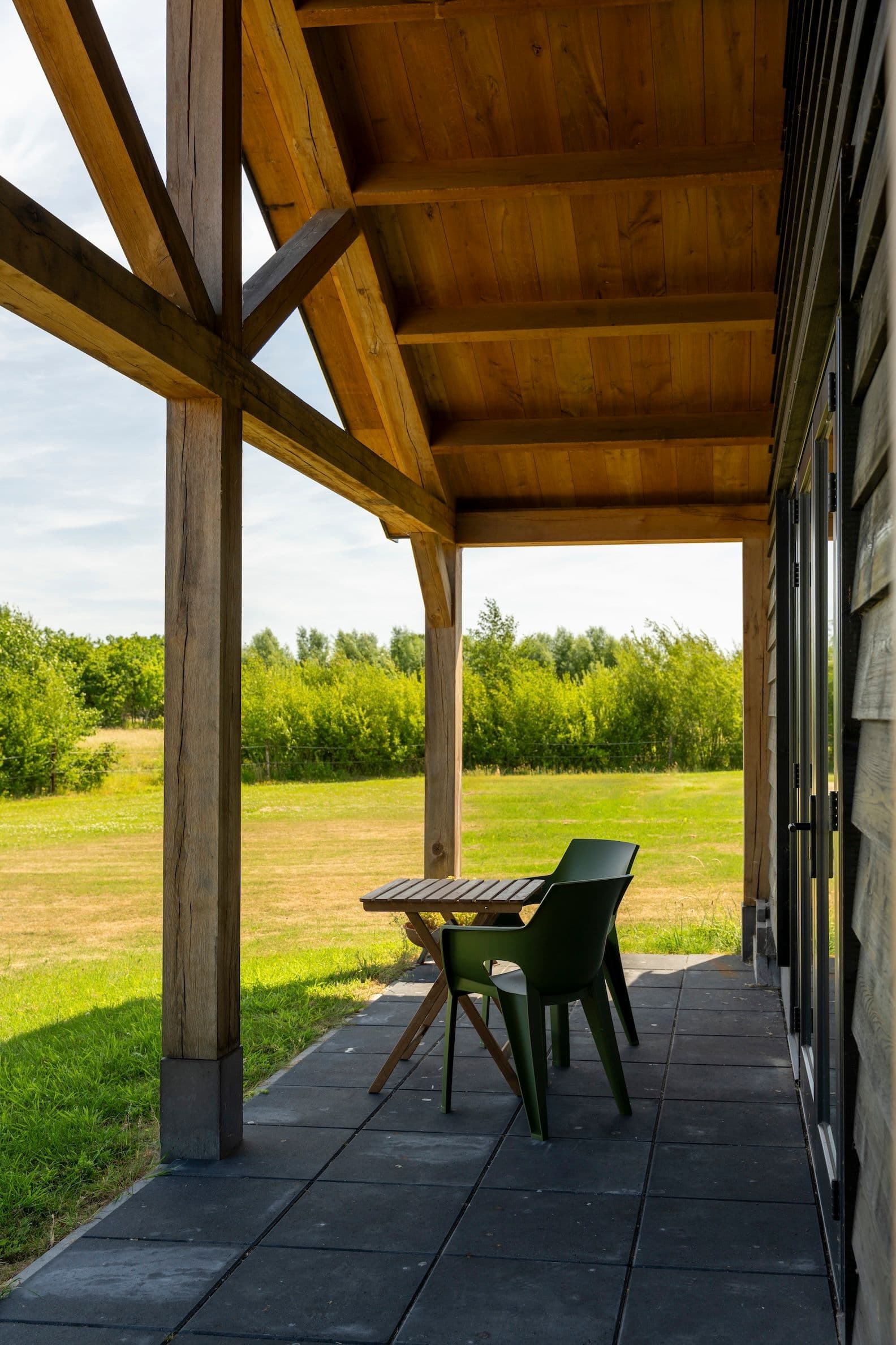 Voorgevel van een overdekt terras met houten balken en plafond, twee groene stoelen en een houten tafel, grasveld en bomen op de achtergrond.