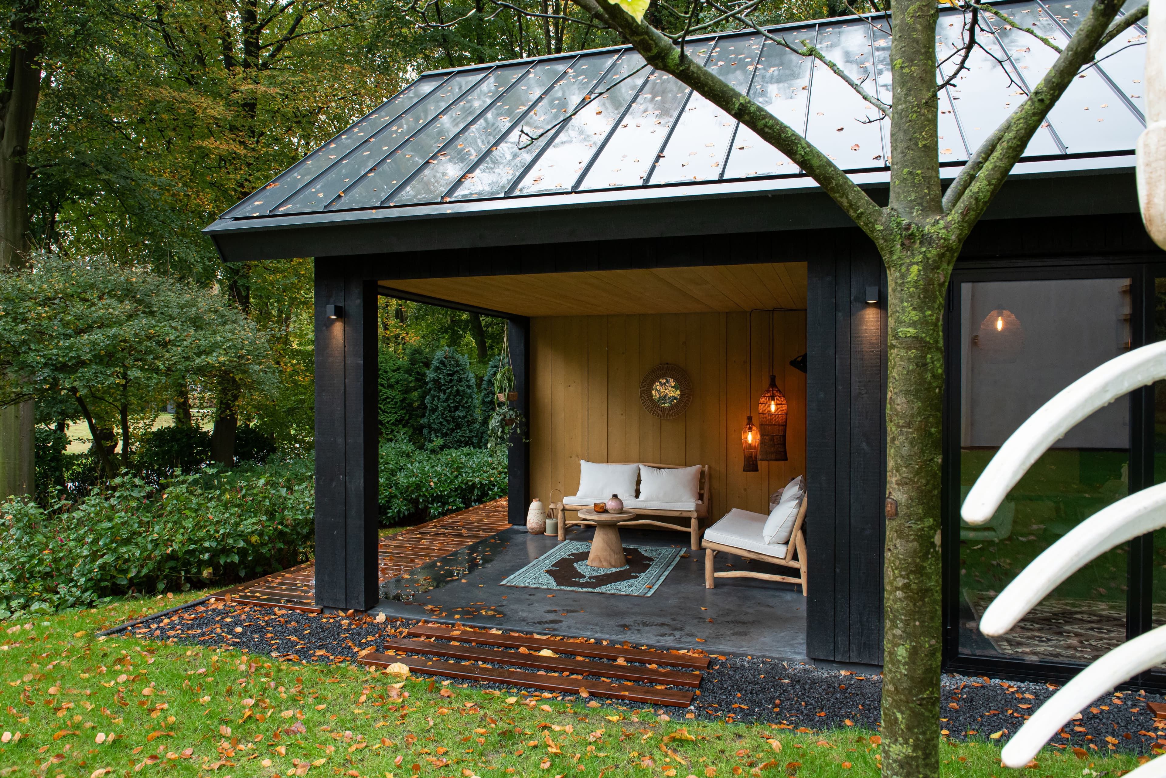 Overdekte zithoek met houten meubels en zachte kussens in een groene tuin met gevallen herfstbladeren