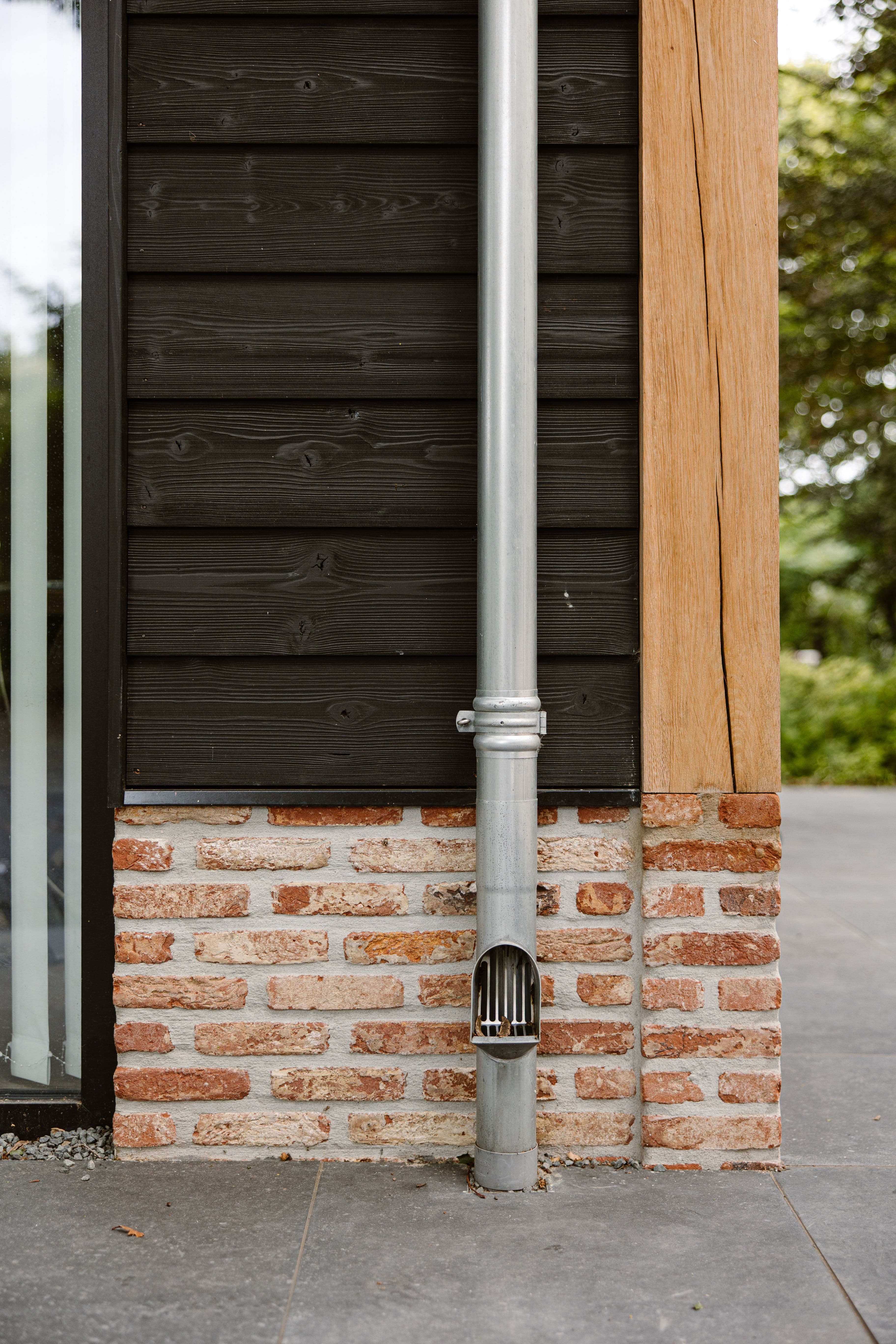 Zilverkleurige regenpijp aan gevel van zwarte houten planken, bakstenen onderzijde en houten balk aan de zijkant