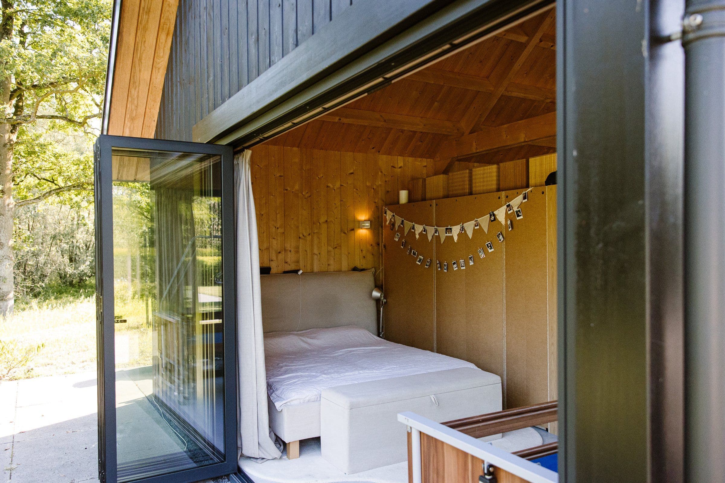 Slaaphoek in een houten buitenhuis met openstaande glazen deur en bed met witte dekens