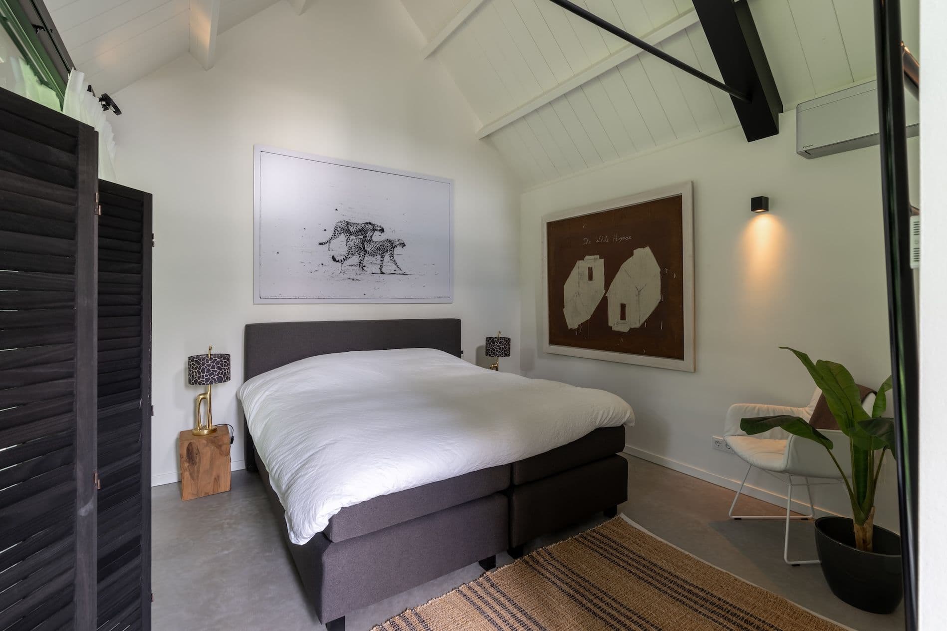 Slaapkamer met een tweepersoonsbed met wit beddengoed, twee schilderijen aan de muur, een witte stoel en een groene plant in een zwarte pot.