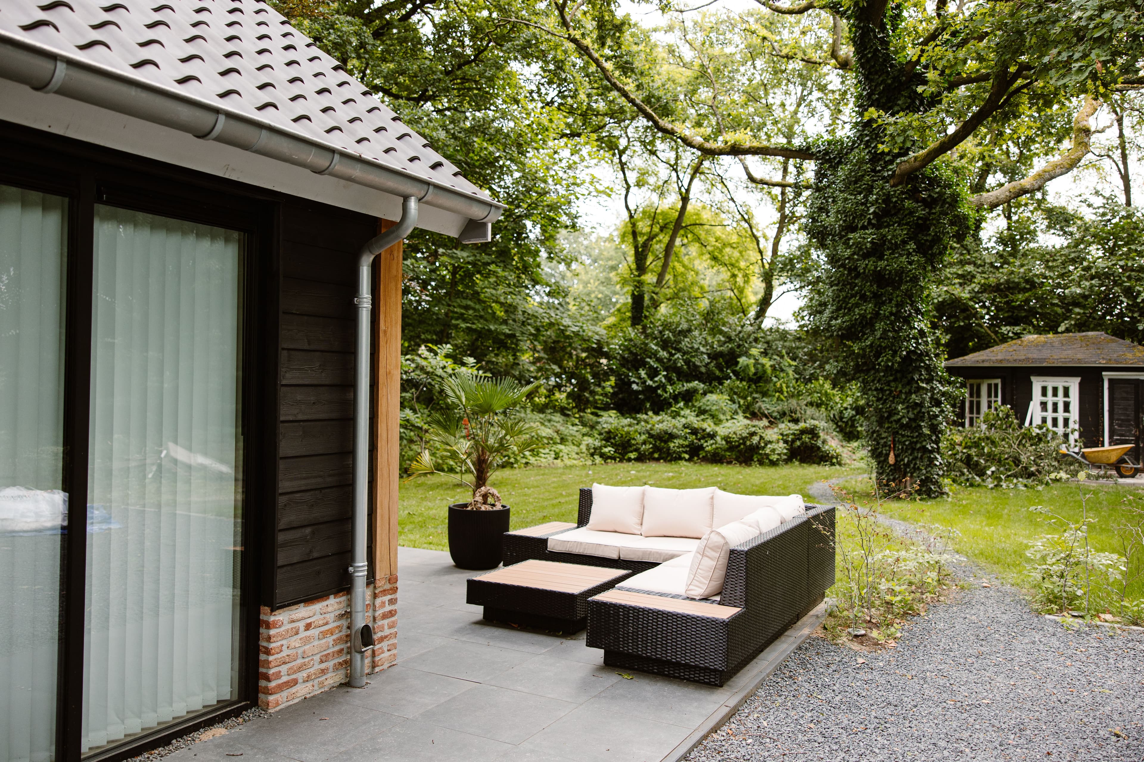 Modern terras met loungebank en planten naast een huis met zwart hout en rode bakstenen, omringd door bomen en groen.