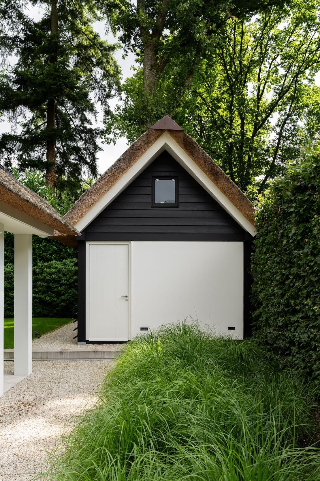 Zwart-wit tuinhuis met rieten dak, witte deur en klein raam, omgeven door groen en siergras langs een grindpad.