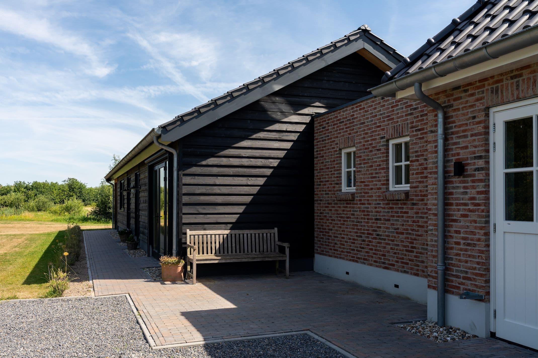 Houten bank staat tegen zwarte houten gevel naast rode bakstenen gevel met kleine ramen en witte deur onder blauwe lucht