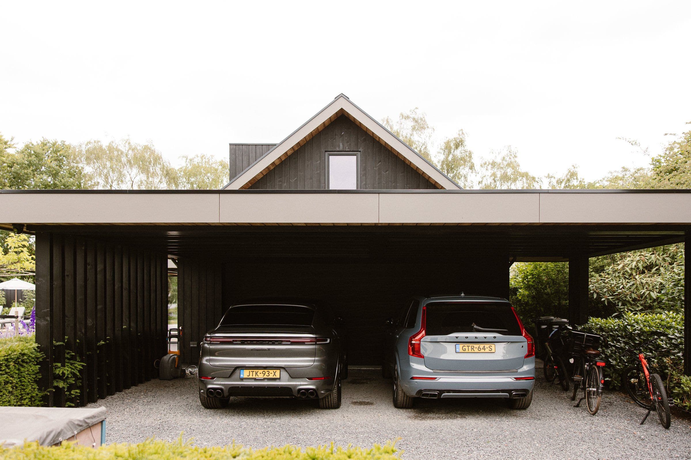 Twee auto's geparkeerd onder een moderne carport bij een huis met zwarte gevel, rechts drie fietsen.