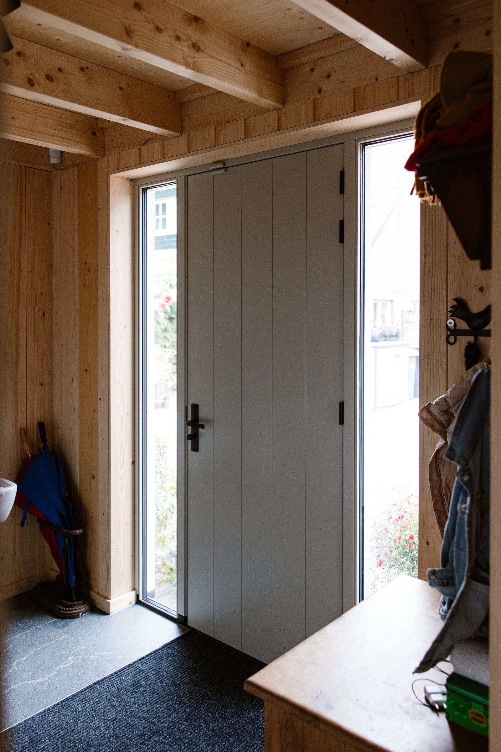 Voordeur van een huis met houten muur, glazen zijpanelen, deurmat en kapstok met jassen