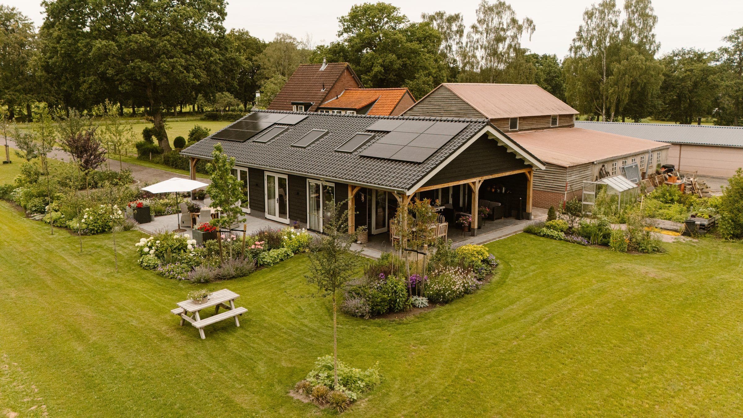 Luchtfoto van een vrijstaand huis met donkere gevel en zonnepanelen op het dak, overdekt terras met zitmeubilair, picnicbank op het gras en kleurrijke tuinborder.