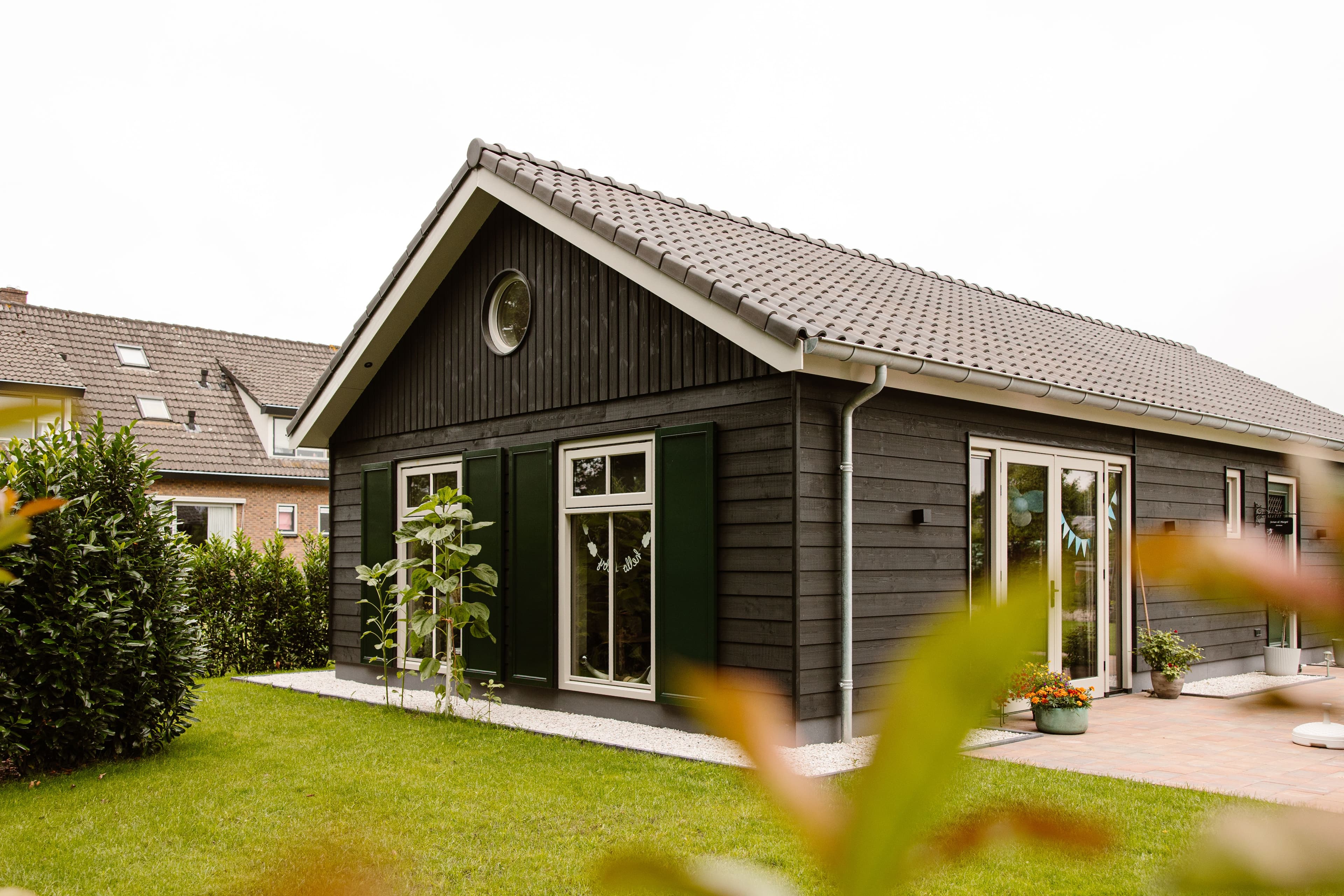 Huis met zwarte houten gevelbekleding en groene luiken, met een goed onderhouden groene tuin en bloemenpotten op het terras.