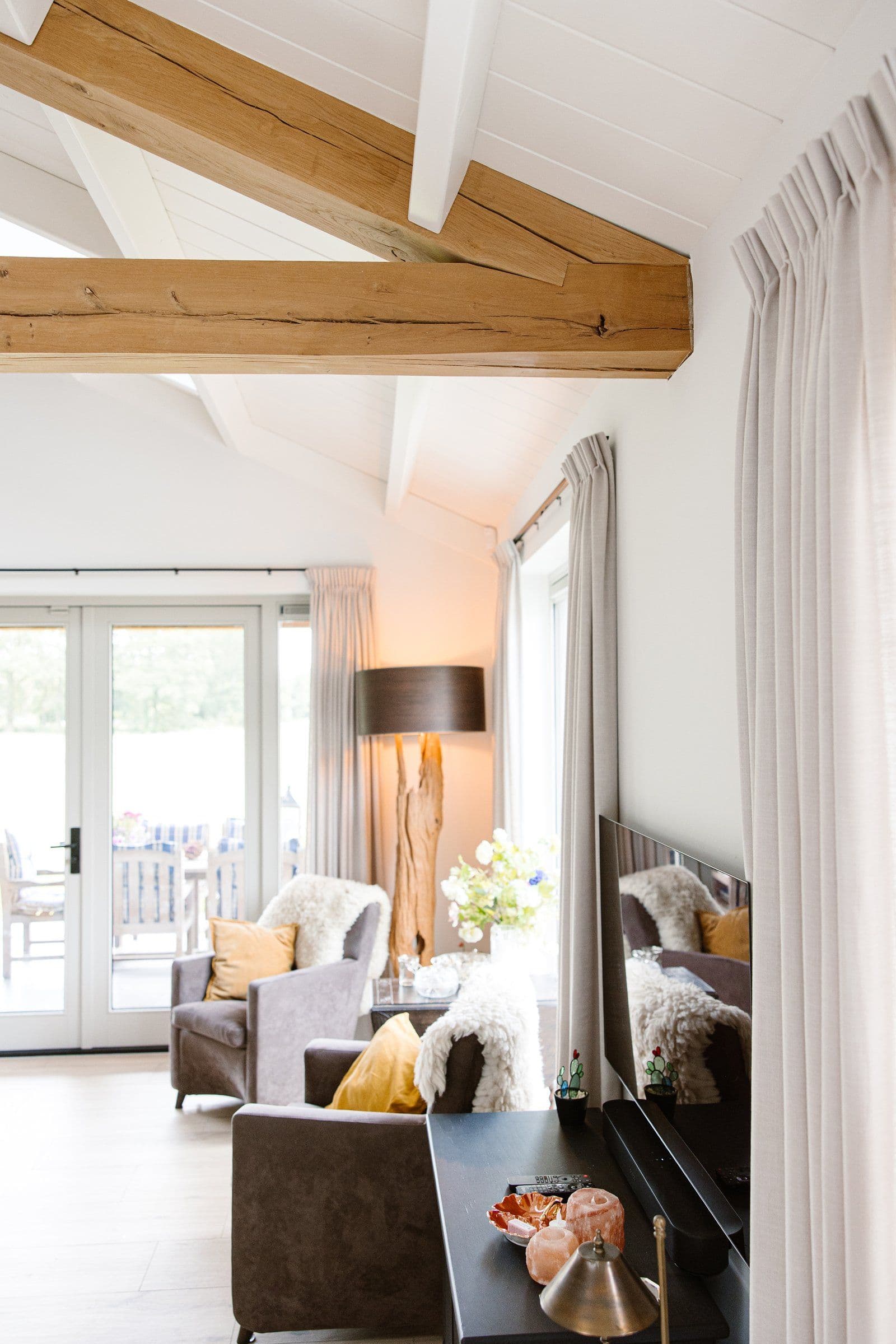 Woonkamer met twee grijze fauteuils bij raampartij, houten plafondbalken en een staande lamp in de hoek