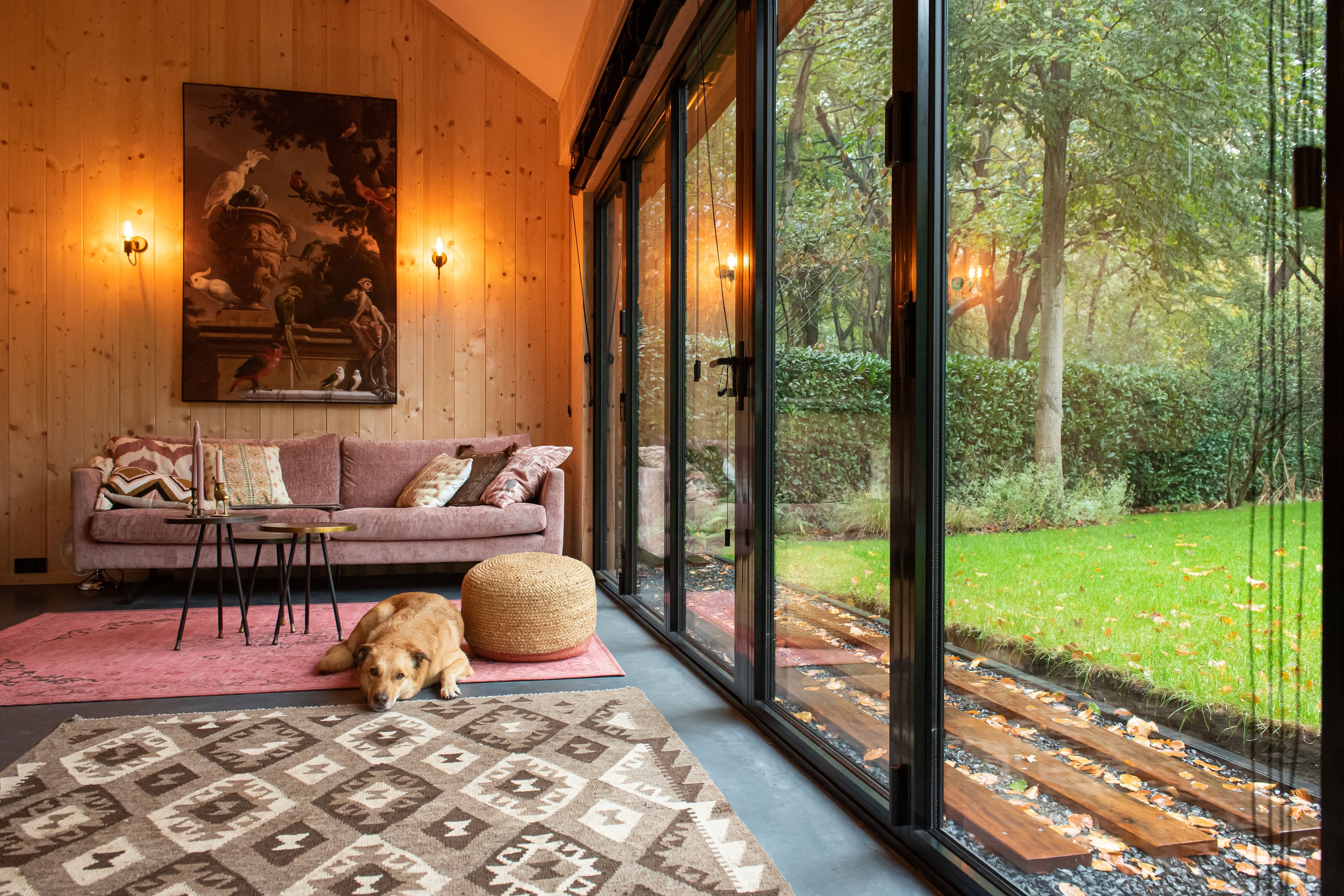 Woonkamer met een roze bank, een hond die op de grond ligt, houten muren en grote glazen schuifdeuren naar een tuin met groene bomen en gras.