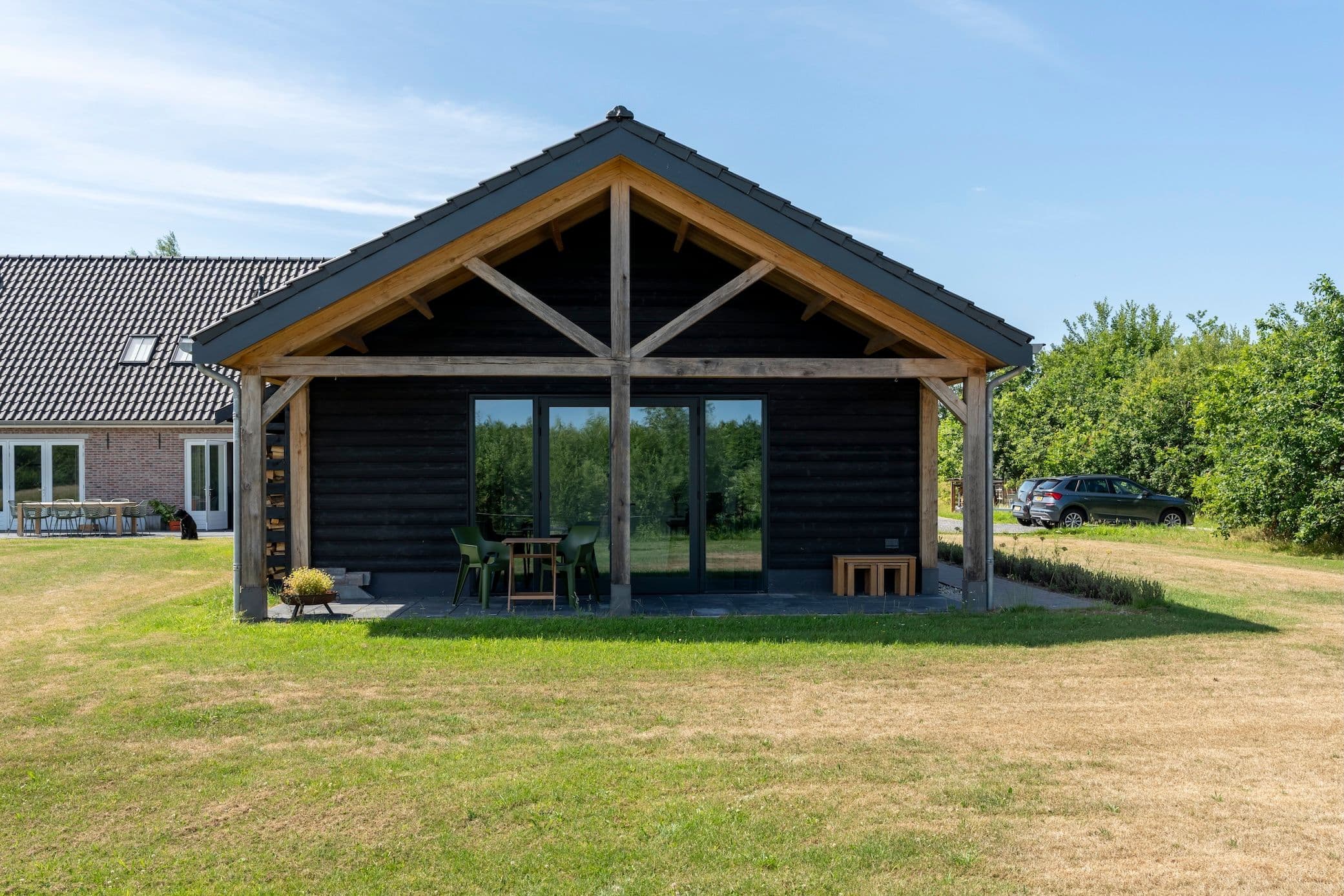 Zwarte houten schuur met houten spanten en glazen schuifdeur, met grasveld op de voorgrond en huis op de achtergrond