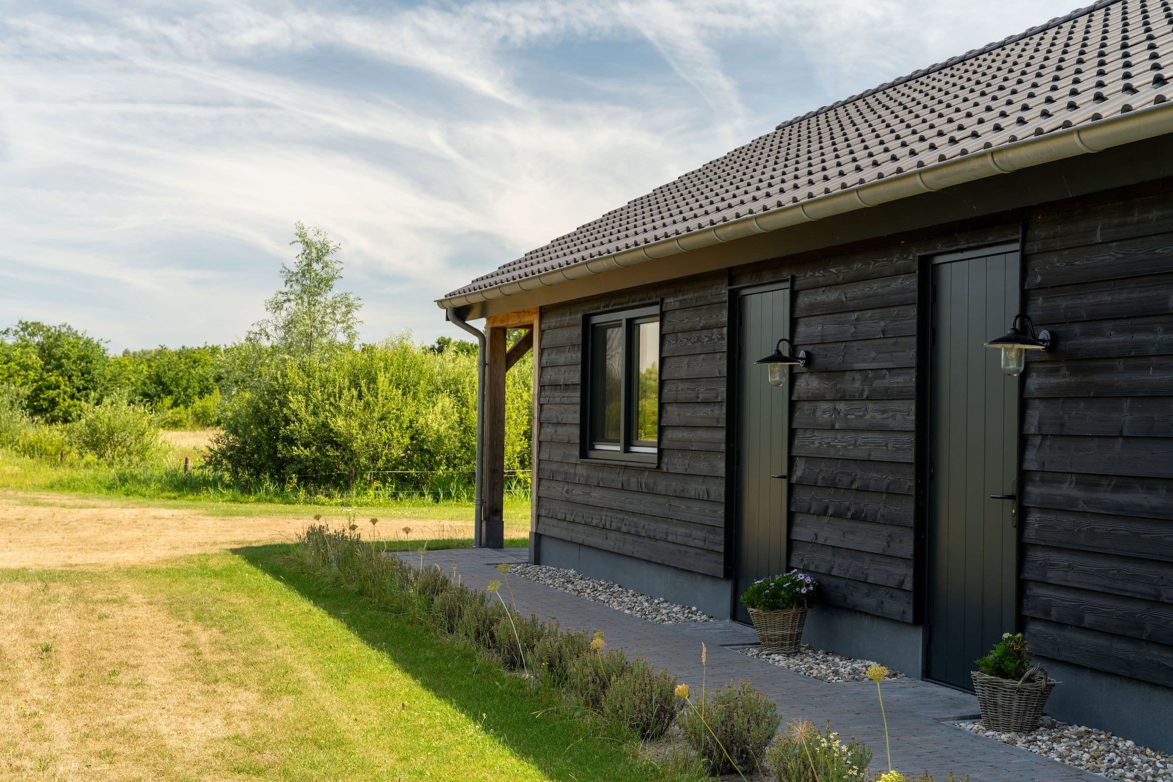 Zijaanzicht van een zwarte houten schuur met twee deuren, een raam en tuin met gras en struiken.
