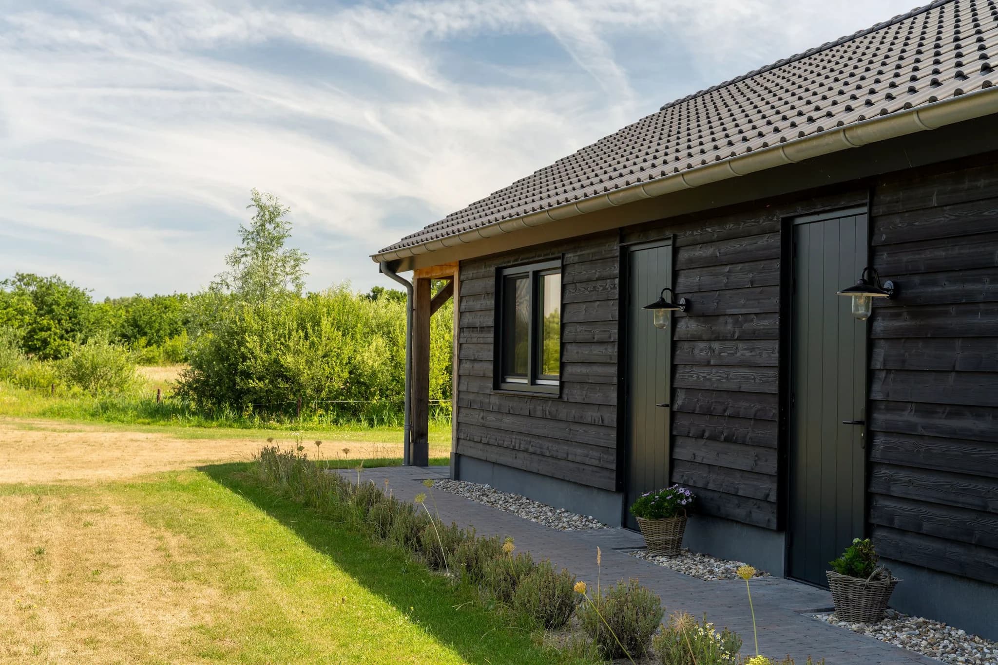 Zijaanzicht van een zwarte houten schuur met twee deuren, een raam en tuin met gras en struiken.