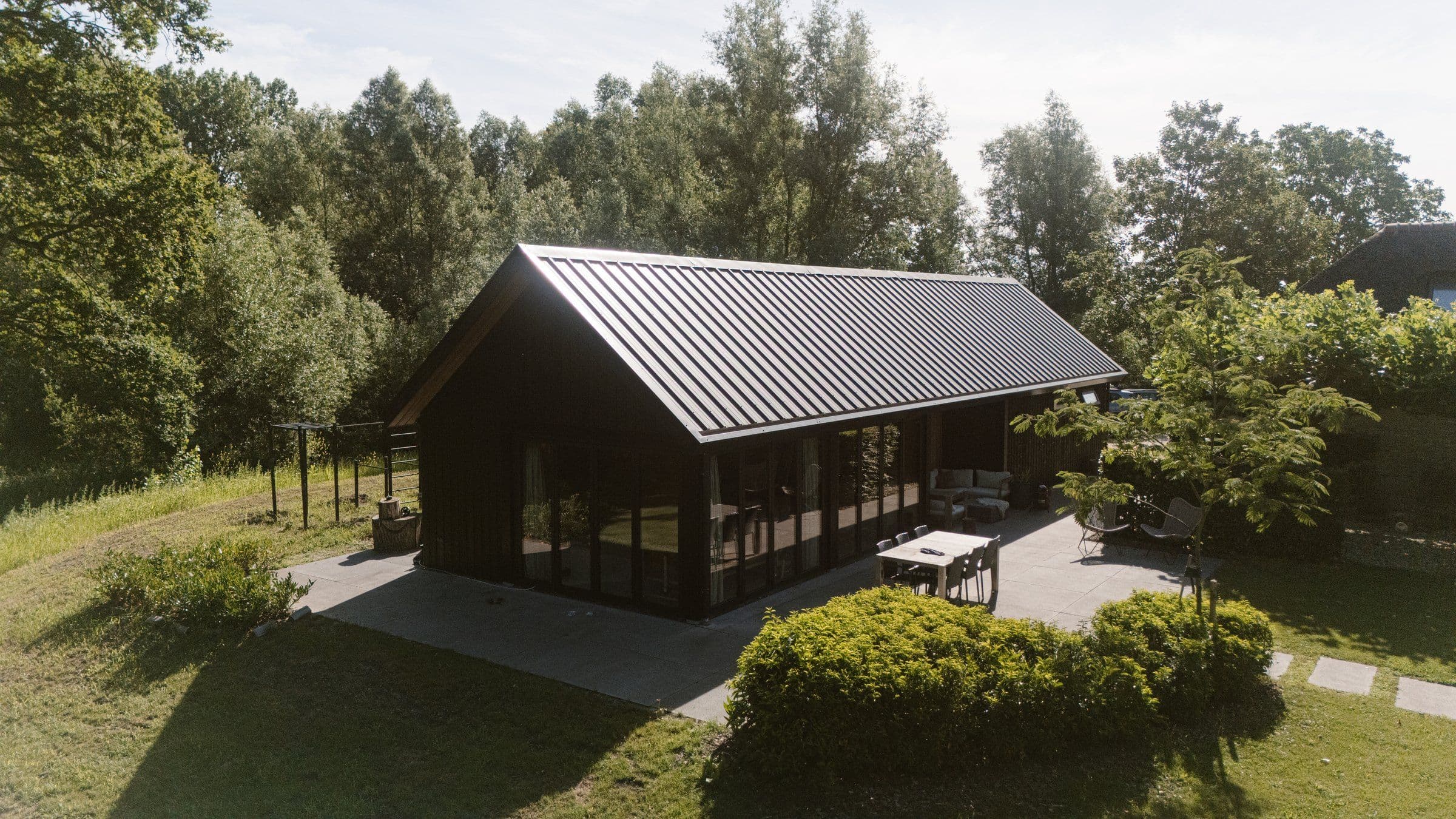 Langwerpige zwarte woning met glazen schuiframen, terras met tafel en stoelen in een groene tuin met bomen en struiken.