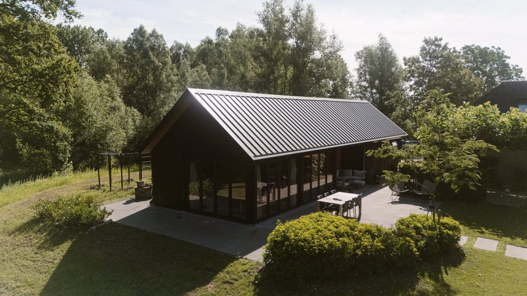 Langwerpige zwarte woning met glazen schuiframen, terras met tafel en stoelen in een groene tuin met bomen en struiken.