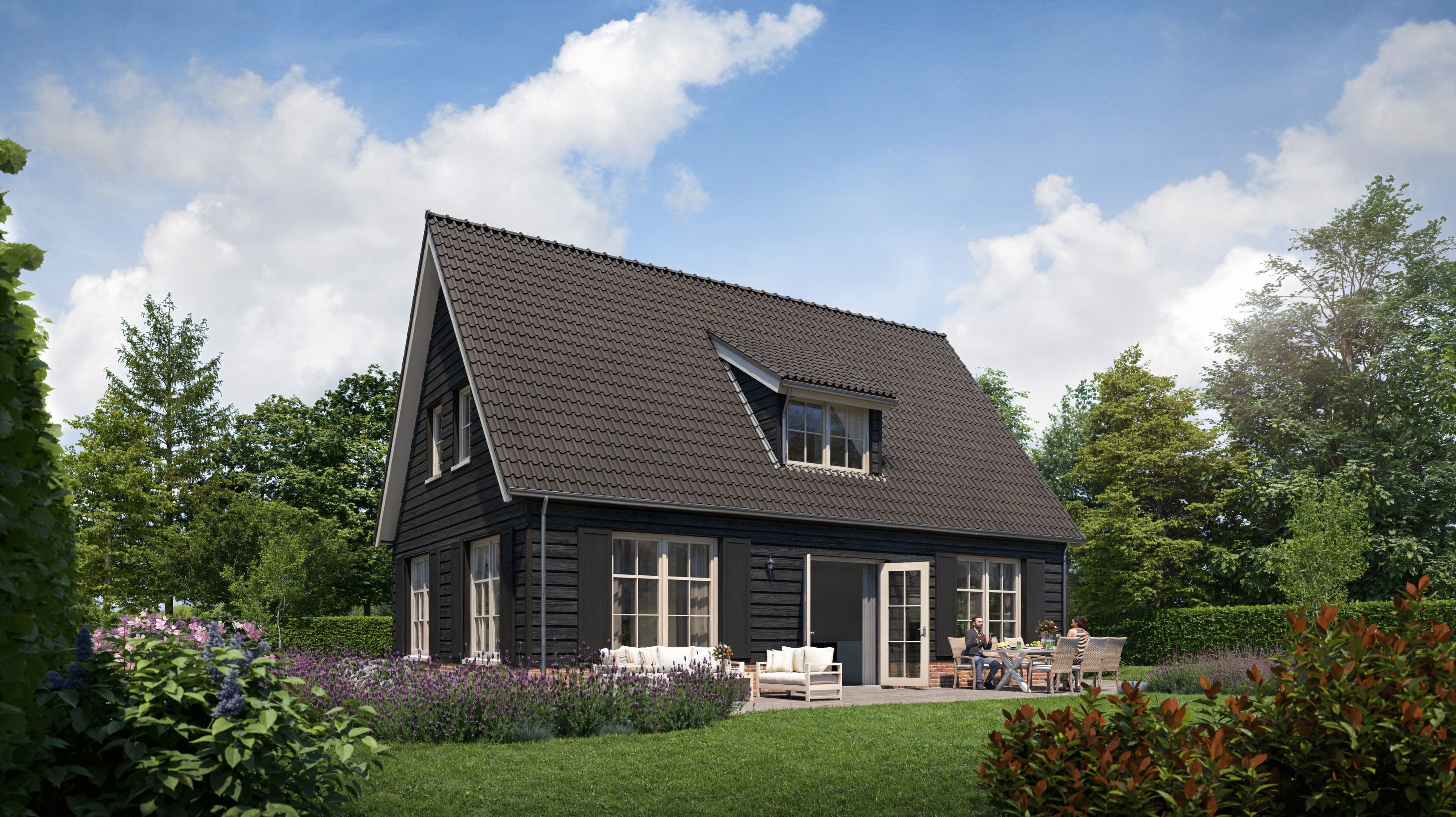 Zwarte houten woning met steil dak en dakkapel, terras met stoelen en tafel en twee personen, omgeven door bloembedden en bomen onder een blauwe lucht.