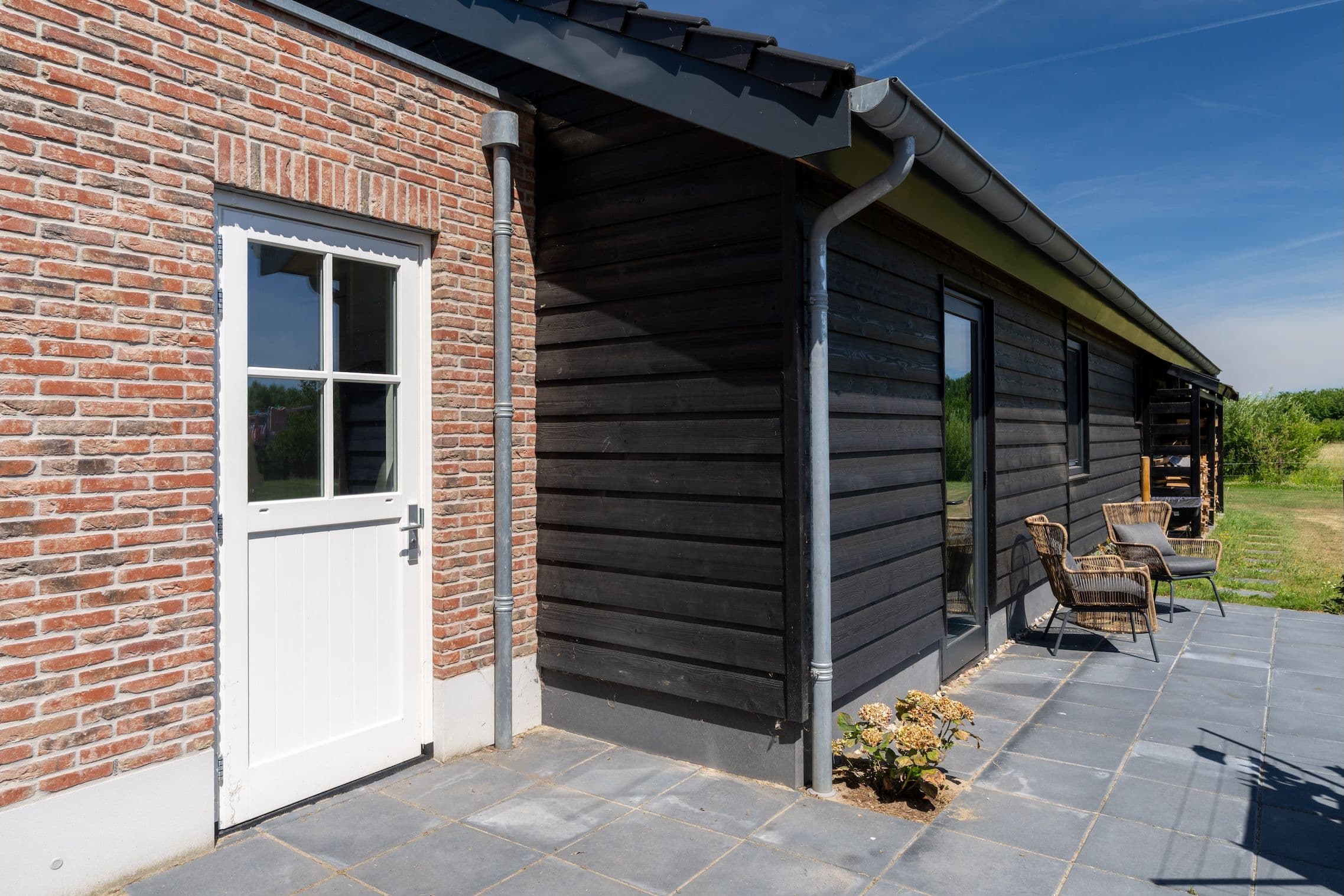 Achterzijde van een huis met een witte deur in bakstenen muur naast een zwarte houten gevel met ramen en twee rieten stoelen op betegeld terras.