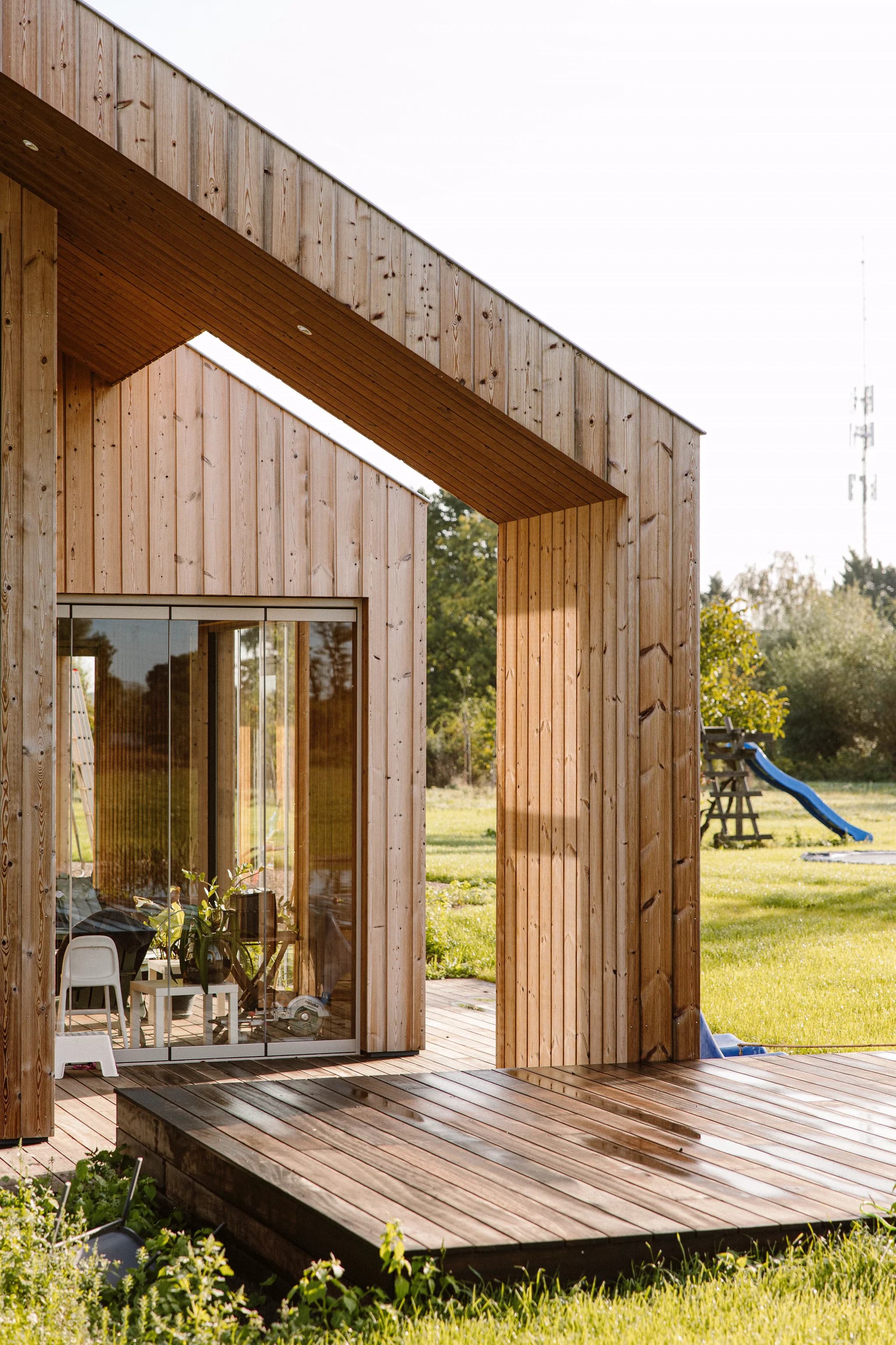 Modern houten huis met glazen schuifdeuren en houten terras in groene tuin met speeltoestel en glijbaan