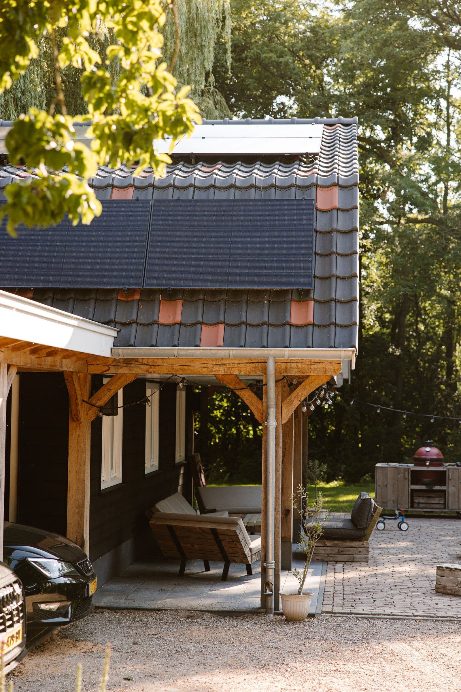 Huis met zwarte dakpannen, zonnepanelen op het dak en houten tuinmeubilair onder een overkapping.