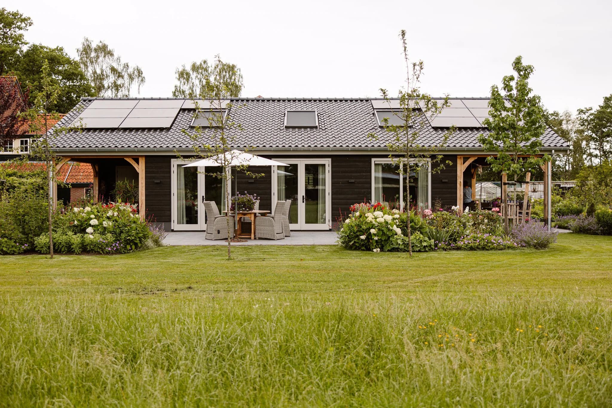 Landelijk huis met zwart houten gevel, zonnepanelen op dak, terras met tuinmeubilair en parasol, omgeven door groene tuin met bloemen en bomen.