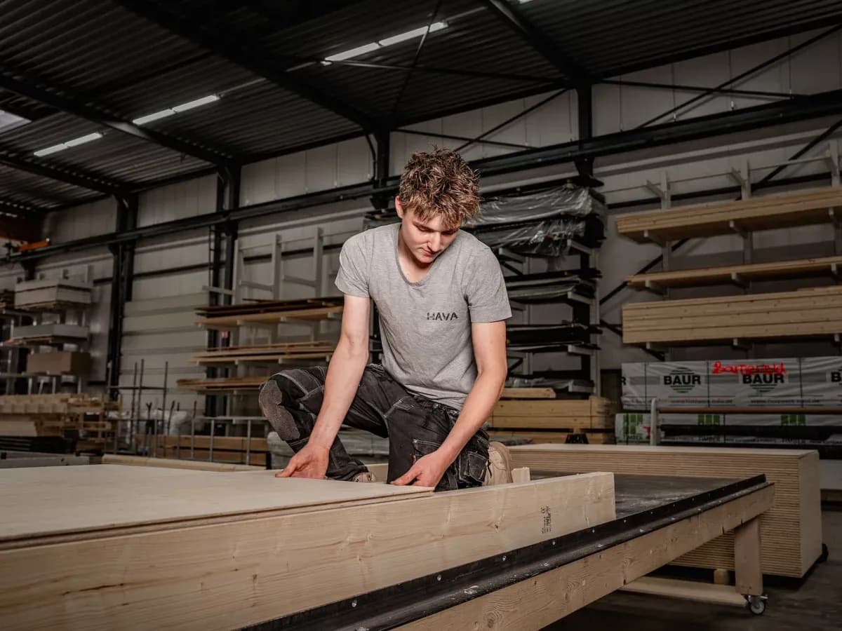 Jonge man in grijs shirt inspecteert houten platen in een opslaghal met houtplanken en rekken