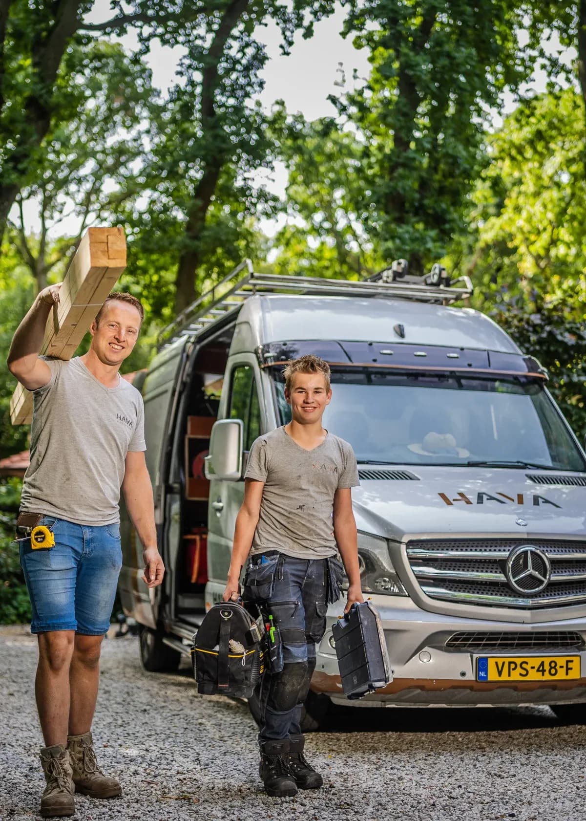 Twee mannen in werkkleding dragen gereedschap en hout terwijl ze voor een grijze bedrijfsbus staan in een groene omgeving