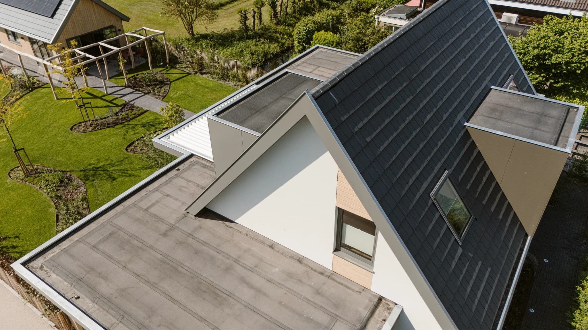 Luchtfoto van een huis met plat dak en schuindak met donkere pannen, omgeven door een tuin met gras en bomen.