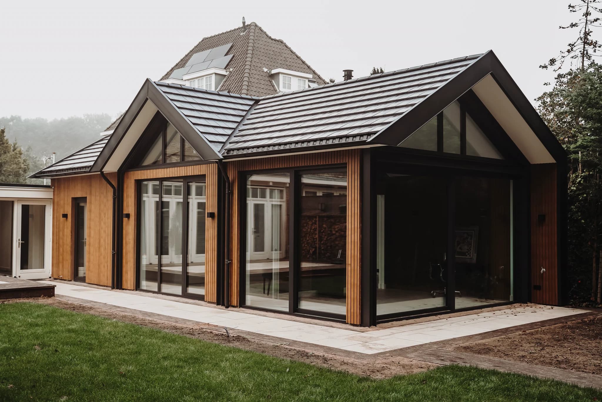 Moderne aanbouw met grote glazen schuifdeuren en zwart pannendak naast een oude woning met zonnepanelen