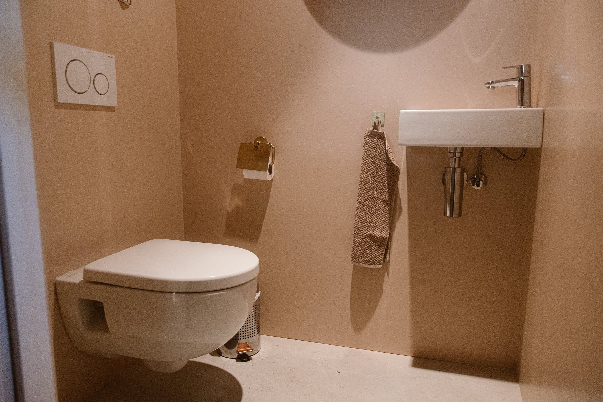 Moderne toiletruimte met hangend wit toilet, klein witte wastafel, beige handdoek aan de muur en een prullenbak op de vloer