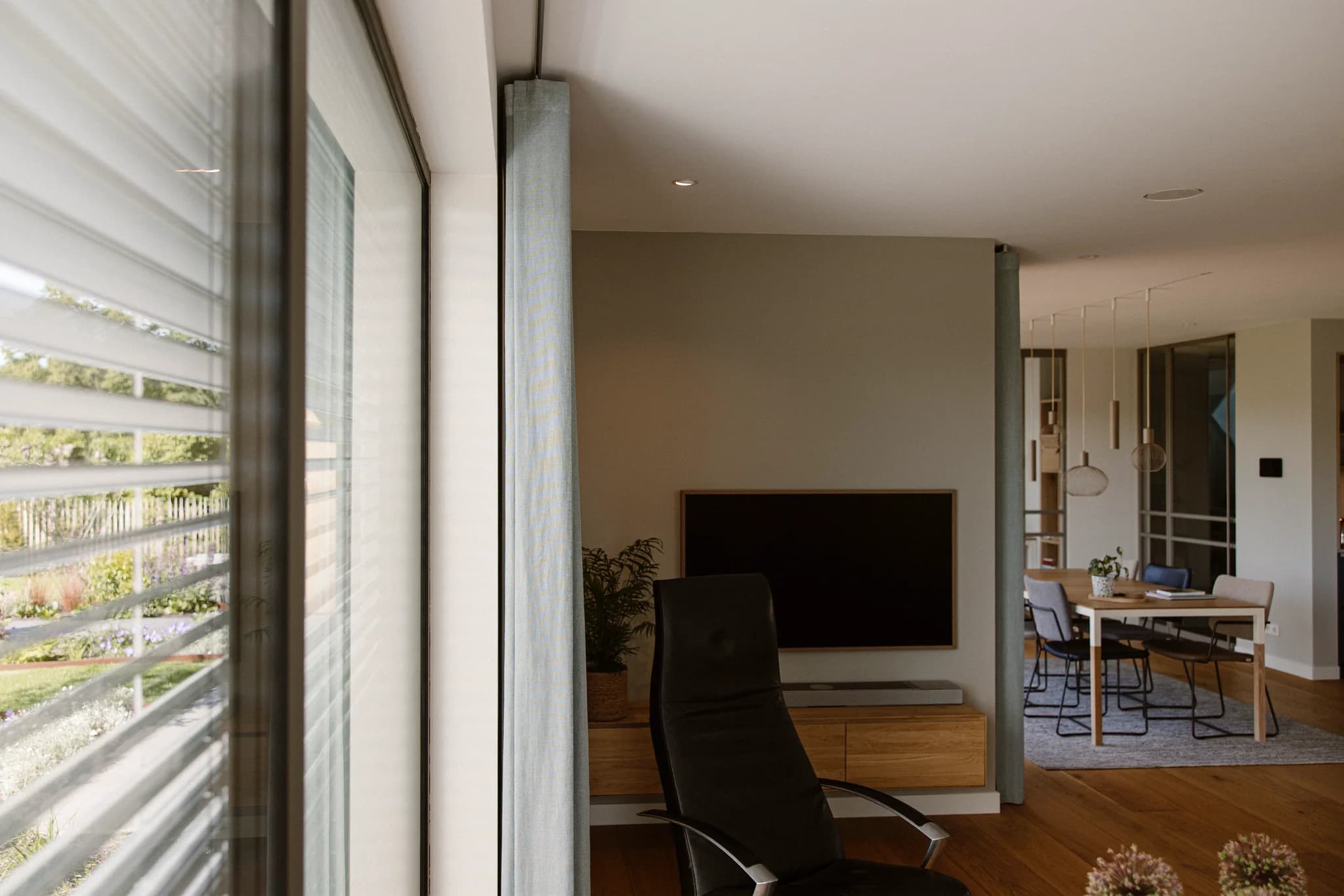 Moderne woonkamer met een zwarte stoel voor een houten meubel met televisie, en in de achtergrond een eettafel met stoelen en hanglampen