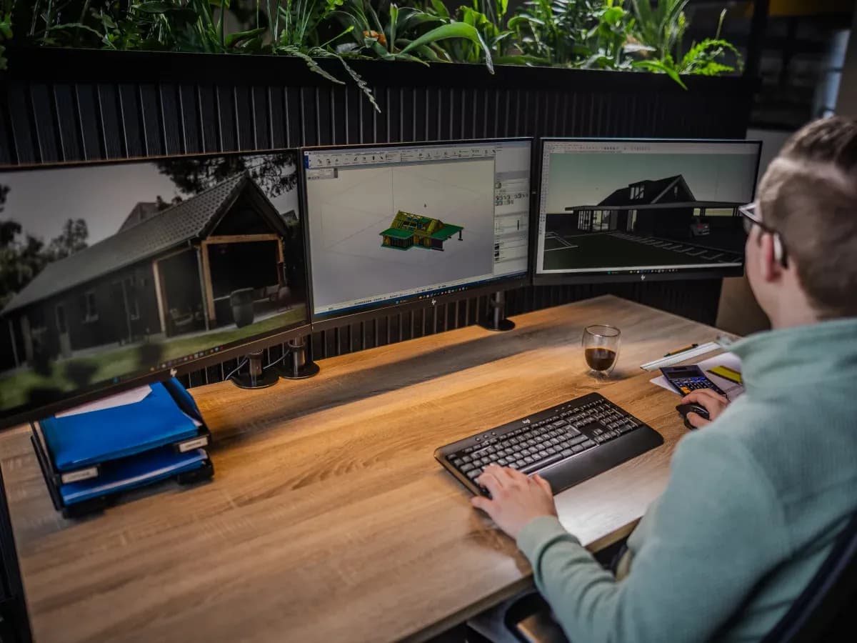 Persoon zit achter bureau en bekijkt ontwerpen van een huis op drie computerschermen