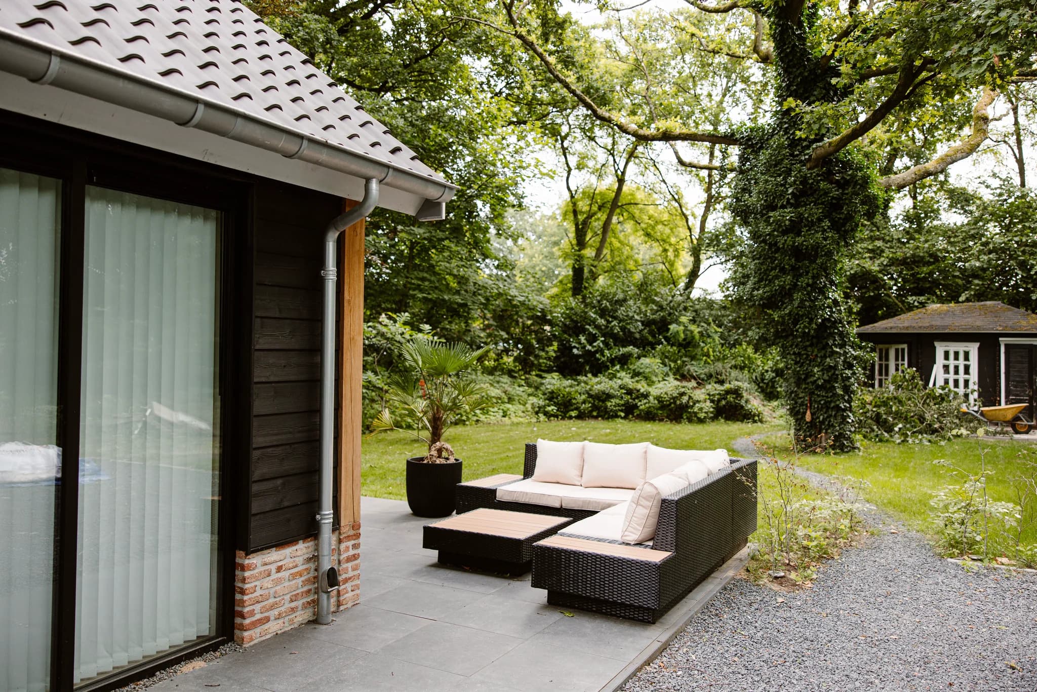 Modern terras met loungebank en planten naast een huis met zwart hout en rode bakstenen, omringd door bomen en groen.