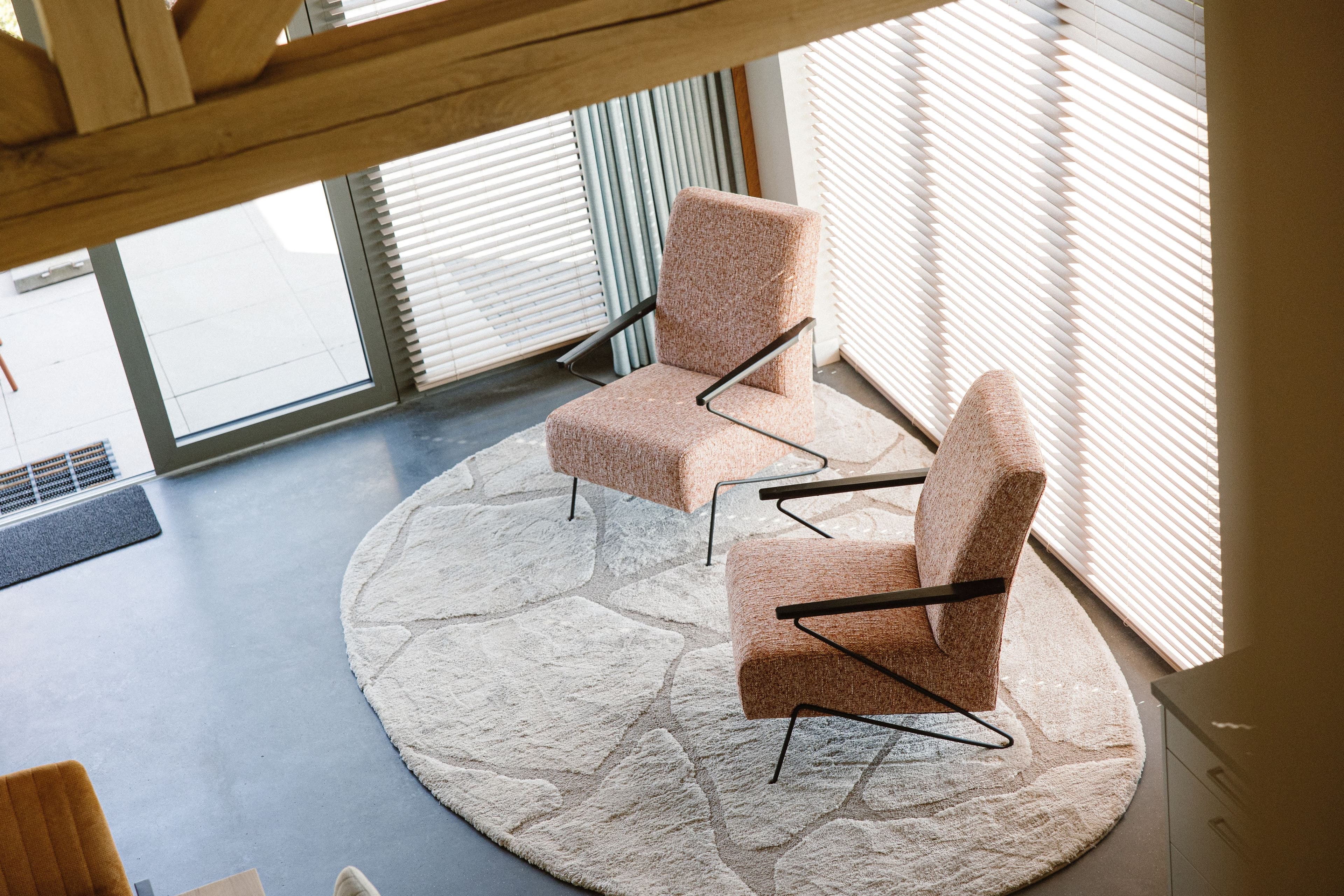 Twee oranjebruine stoffen stoelen met metalen armleuningen op een rond beige vloerkleed bij een raam met jaloezieën