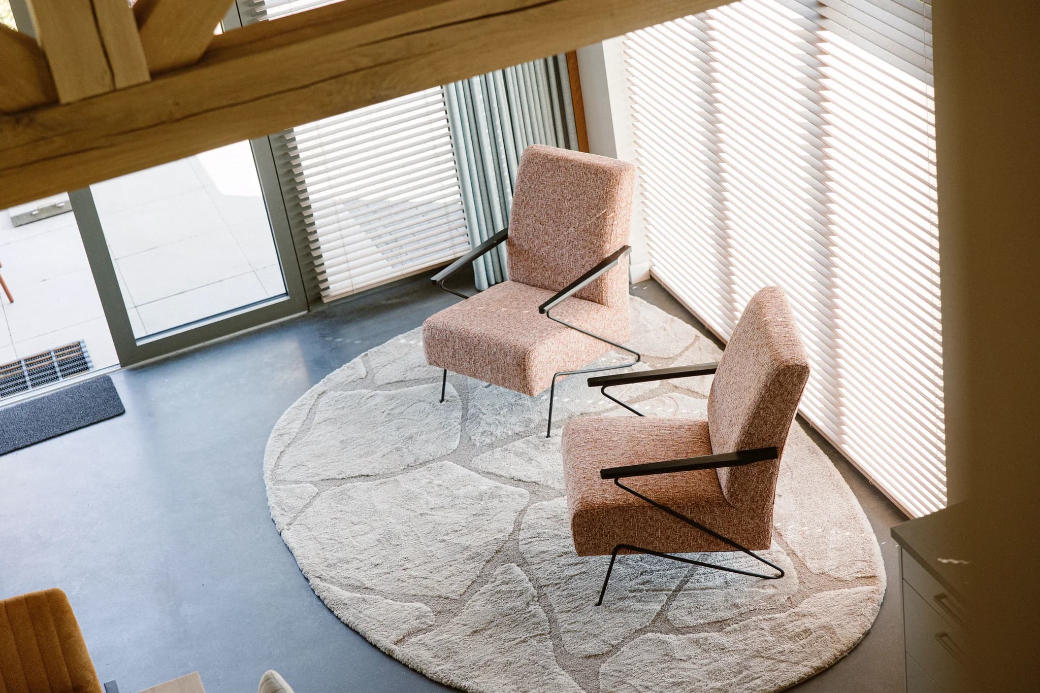 Twee oranjebruine stoffen stoelen met metalen armleuningen op een rond beige vloerkleed bij een raam met jaloezieën